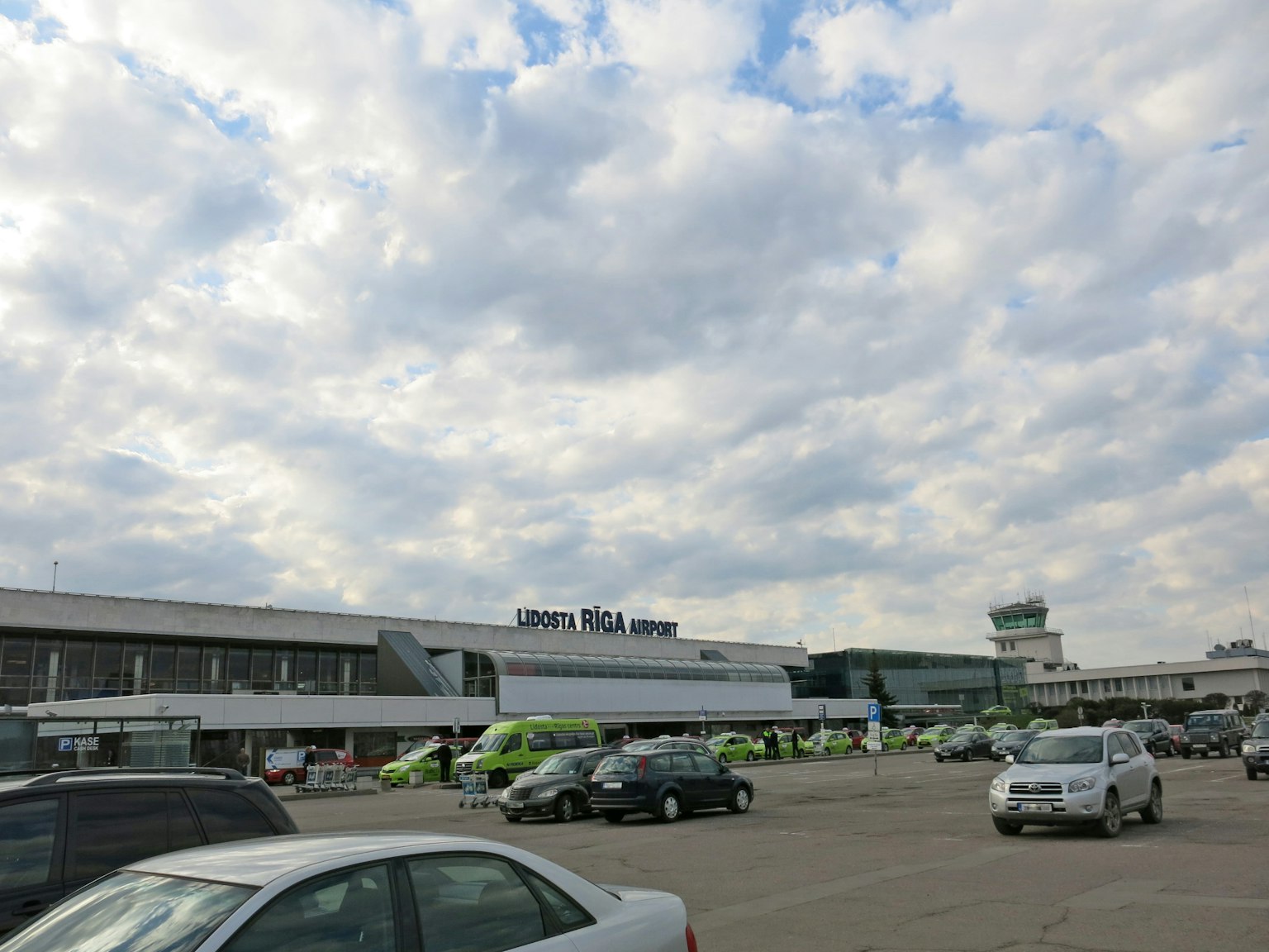 Vue d'un bâtiment d'aéroport et d'un parking sous un ciel nuageux avec des véhicules garés