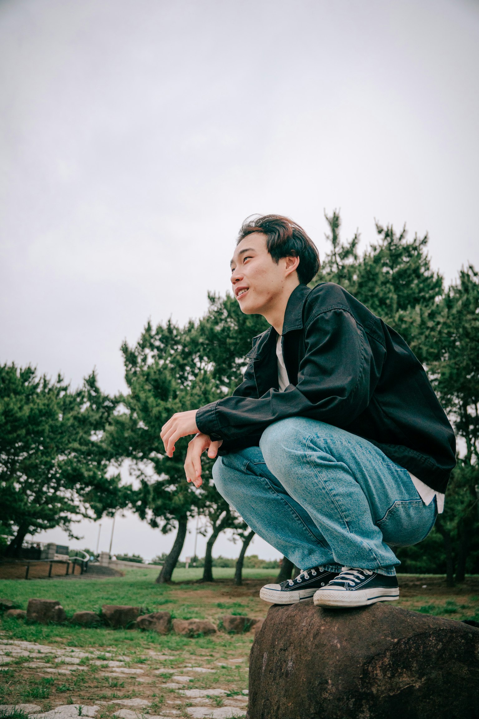 Young man squatting on a stone in a park surrounded by green trees and cloudy sky