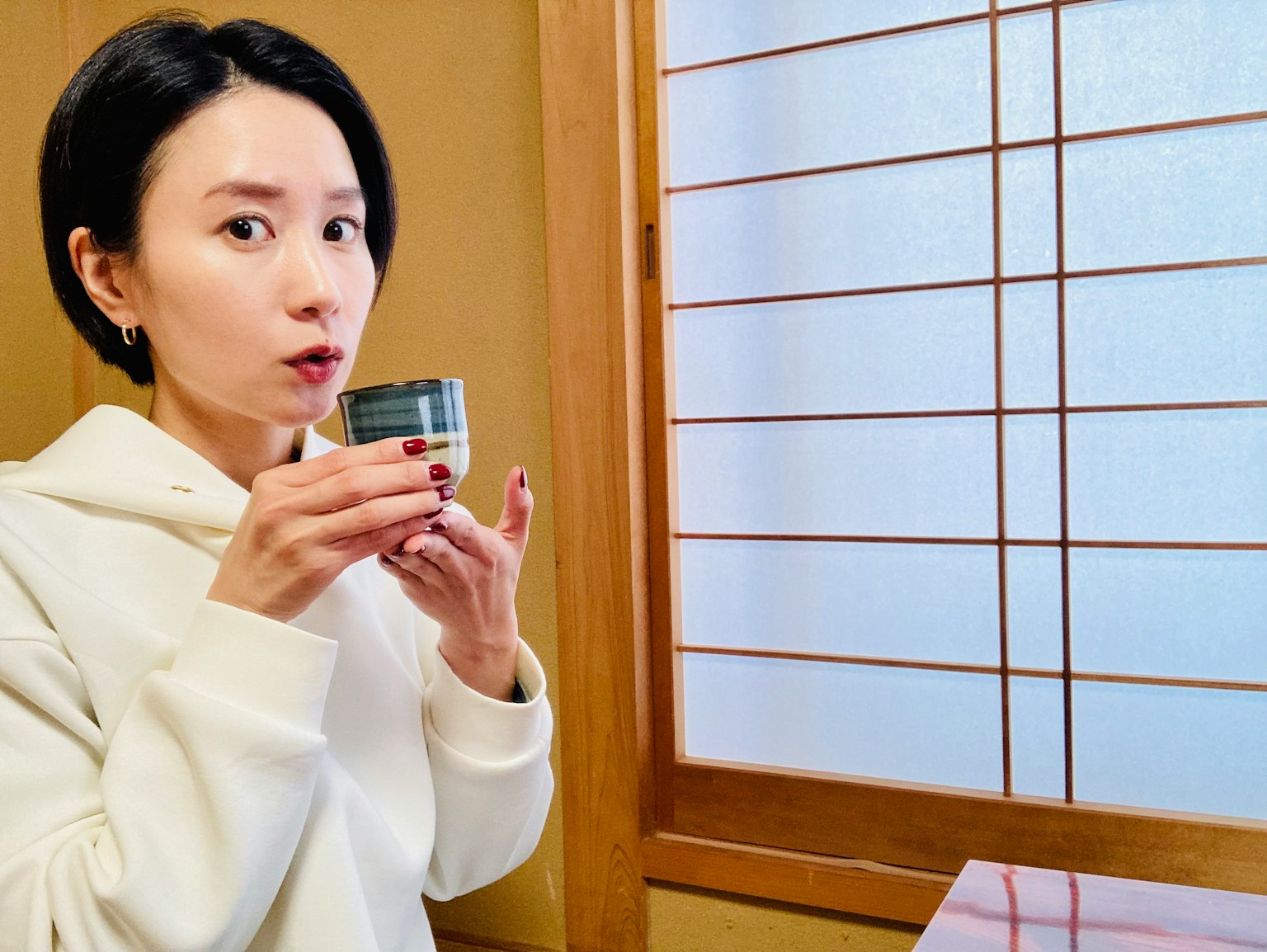 A woman in a white hoodie holding a small cup near a window