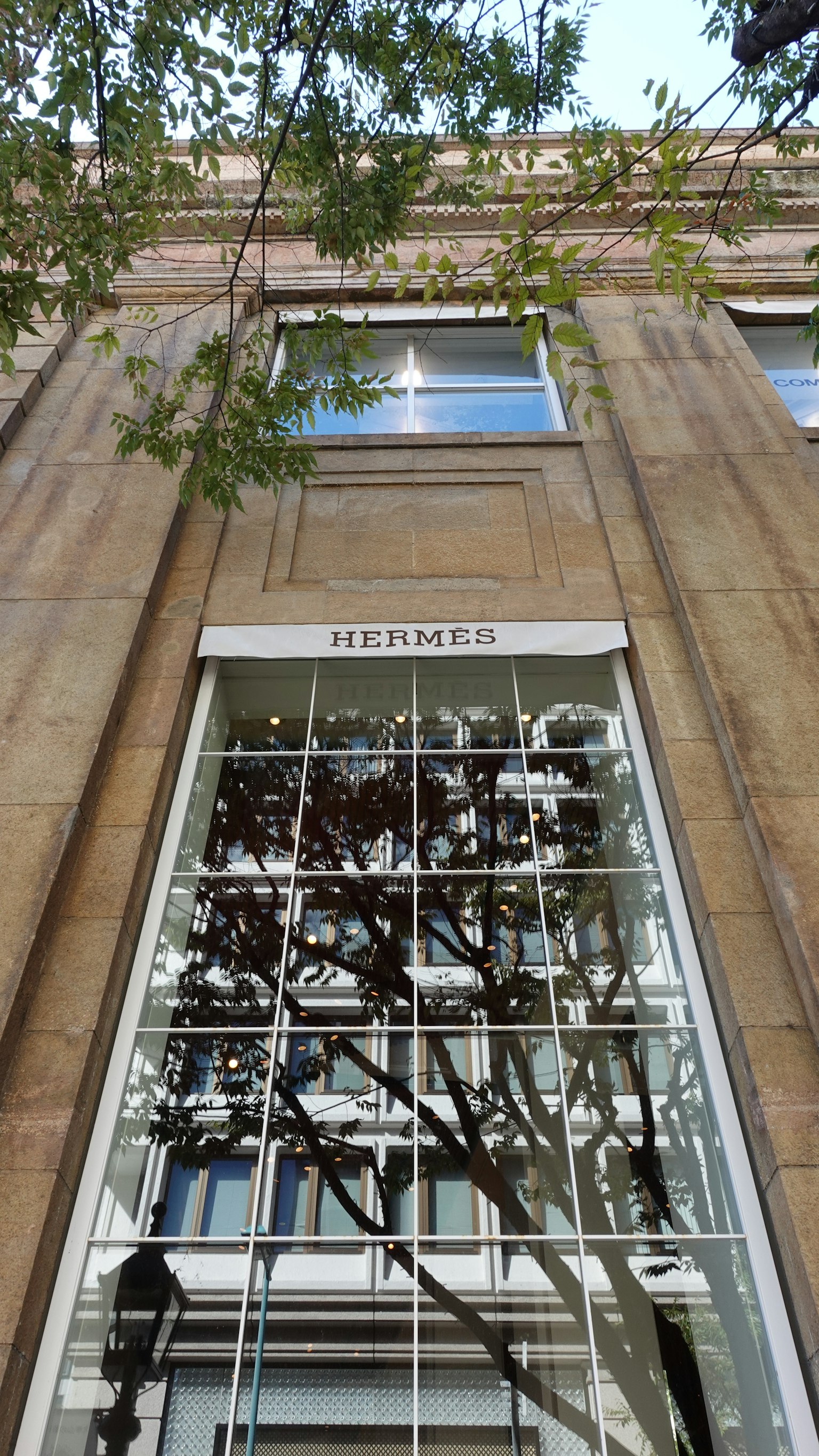 ภาพภายนอกของร้าน Hermès ที่มีหน้าต่างกระจกขนาดใหญ่และเงาของต้นไม้