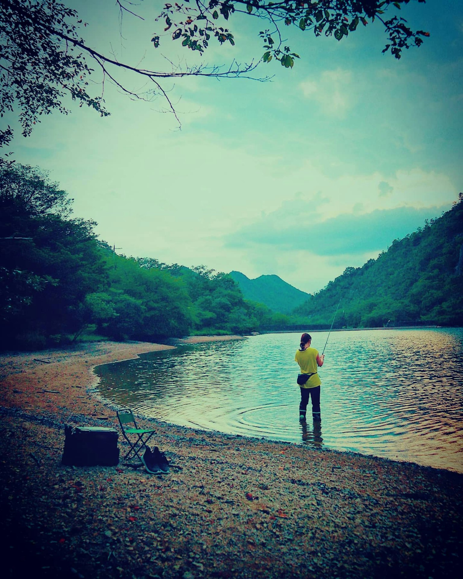 Một người đang câu cá bên bờ sông, xung quanh là những ngọn núi xanh tươi và mặt nước yên tĩnh
