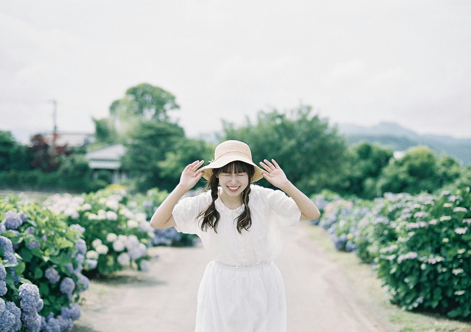 A woman in a white dress and hat is smiling and posing surrounded by blue hydrangeas