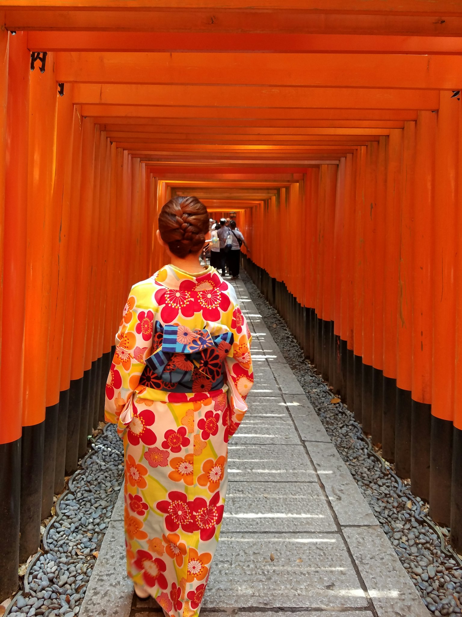 Donna in kimono floreale che cammina attraverso un tunnel di torii arancioni