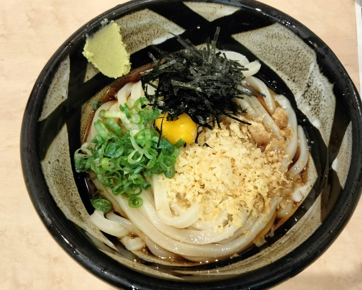 Fideos udon servidos en un tazón negro cubiertos con cebollín, yema de huevo, algas y copos de tempura