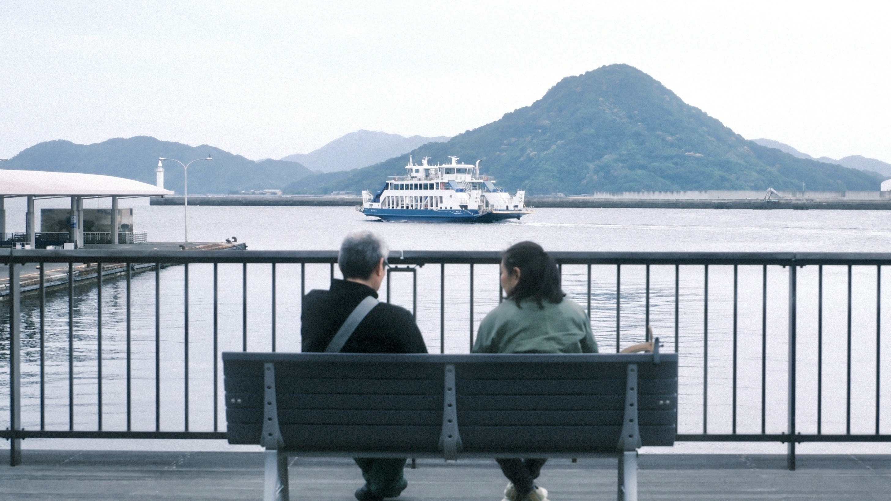 海辺の遊歩道でベンチに座る人とフェリー