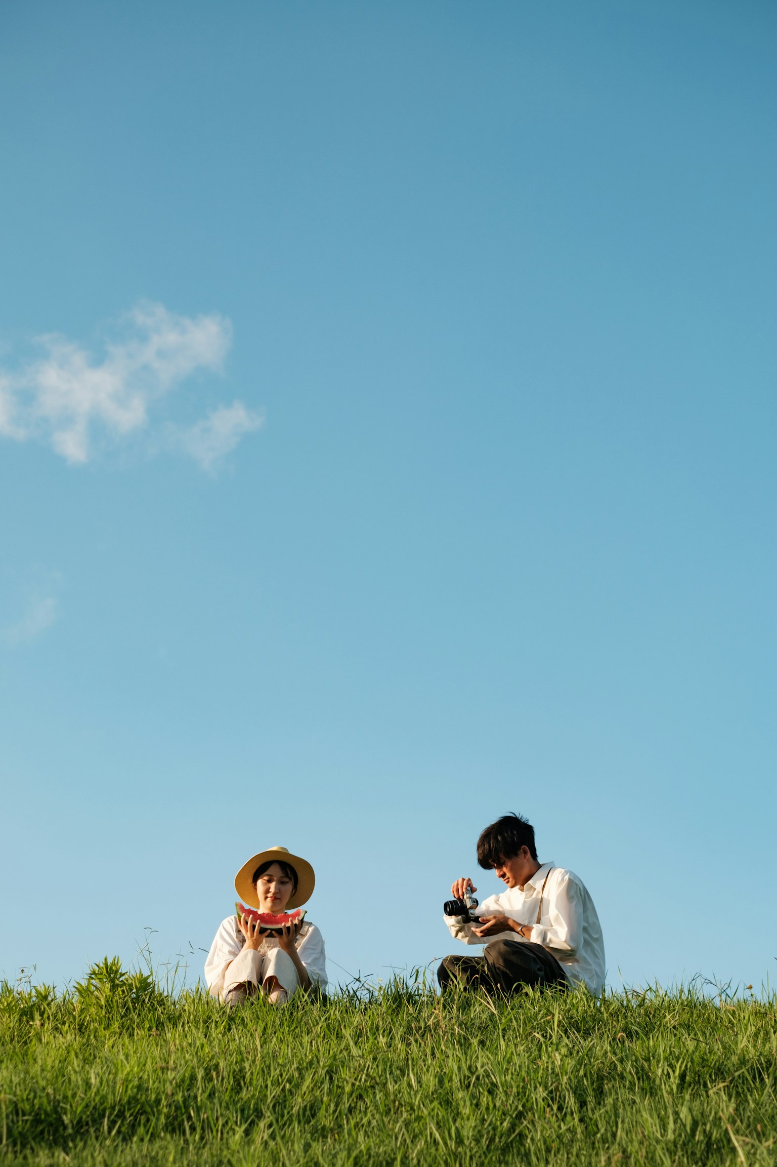 Dua orang duduk di atas rumput di bawah langit biru Satu memegang kamera sementara yang lain bersantai