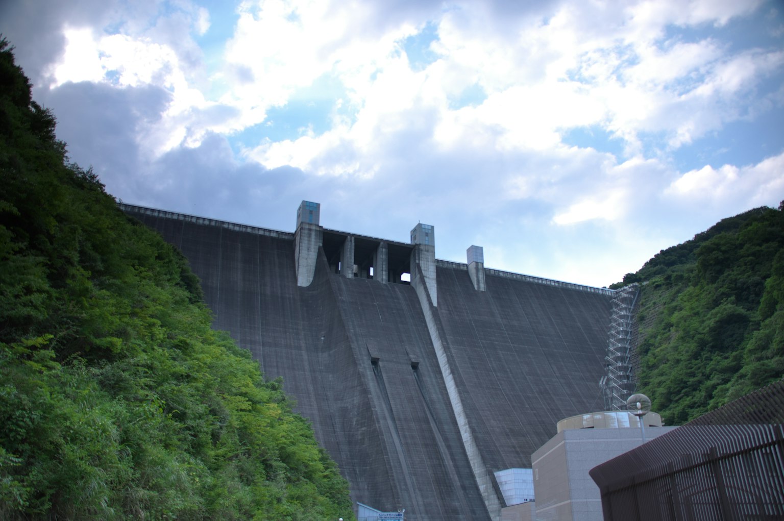 A massive concrete dam surrounded by green hills under a cloudy sky