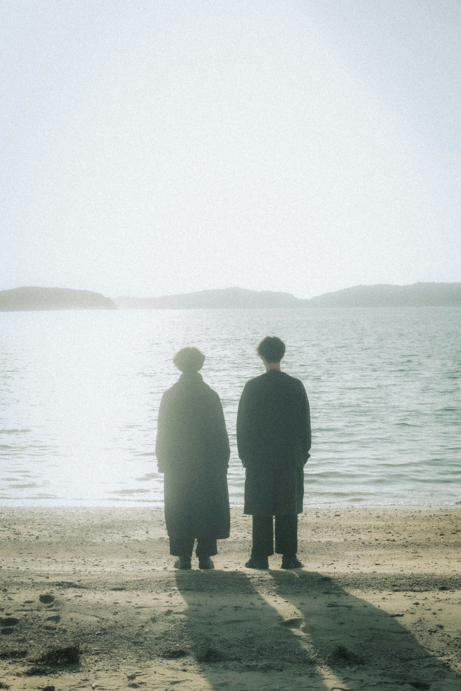 Two figures standing on a beach silhouetted against the water sunlight reflecting on the surface
