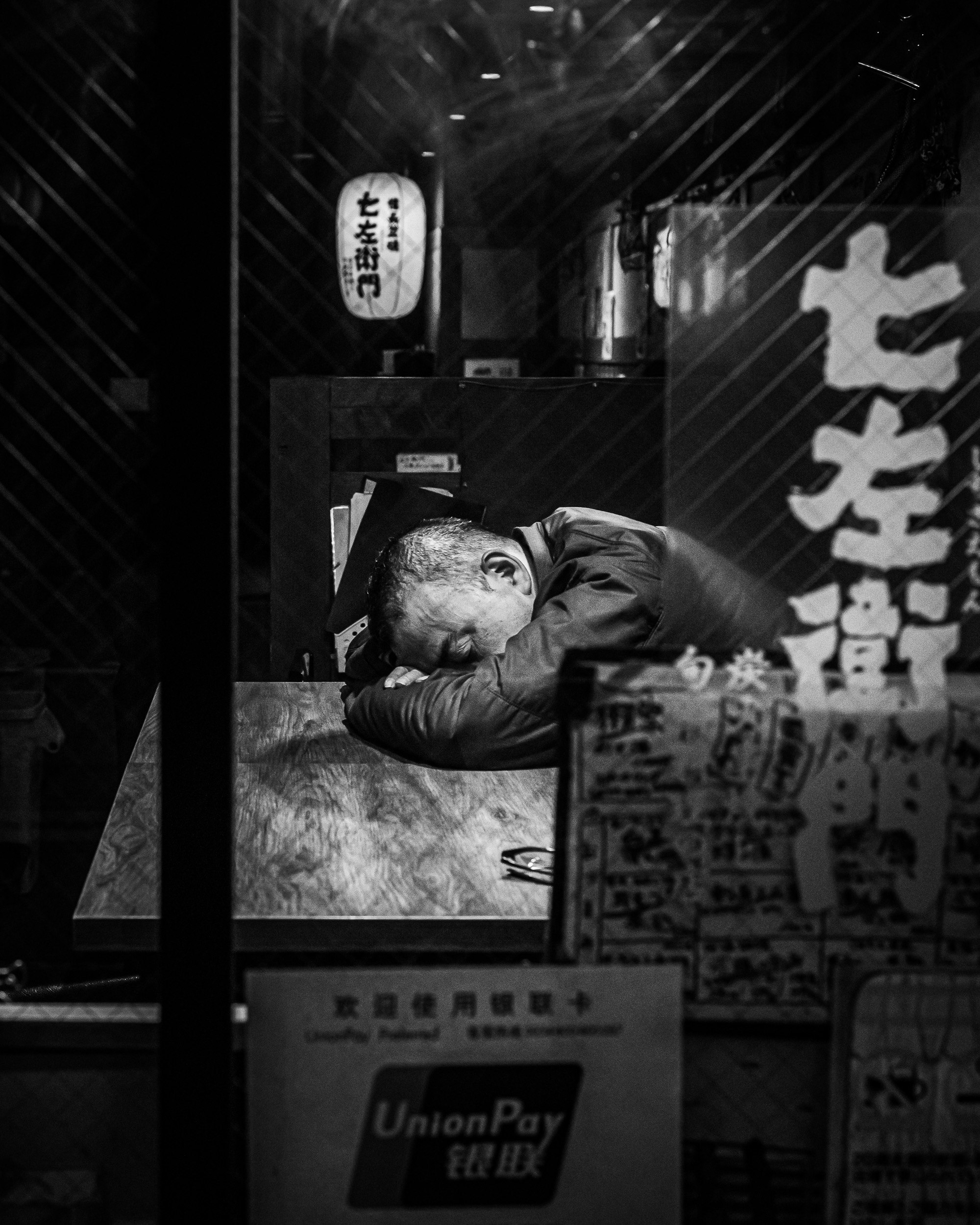黒白の写真で、男性がテーブルに頭を伏せている様子が描かれている