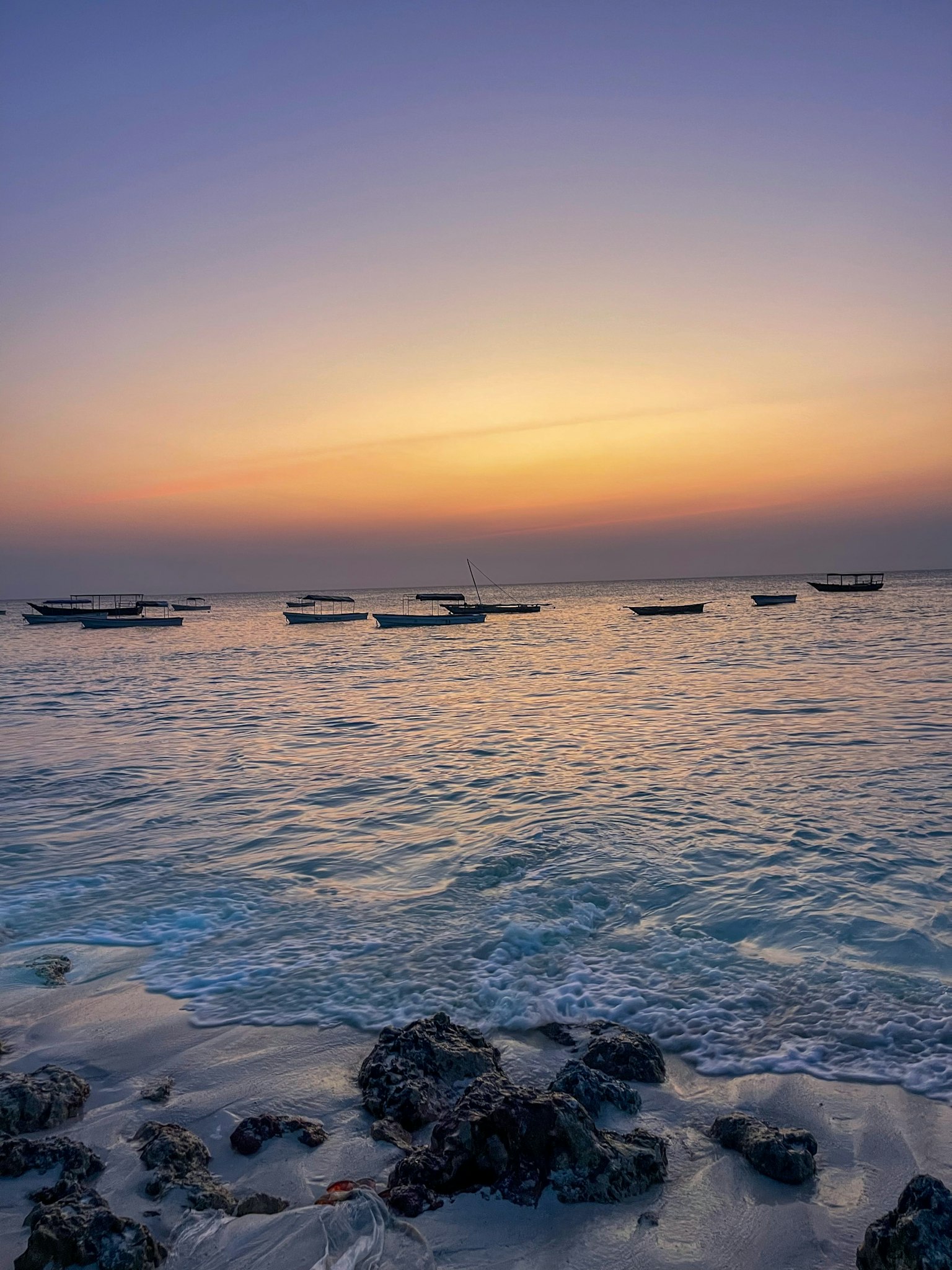 Paisaje marino sereno con atardecer y barcos flotantes costa rocosa en primer plano