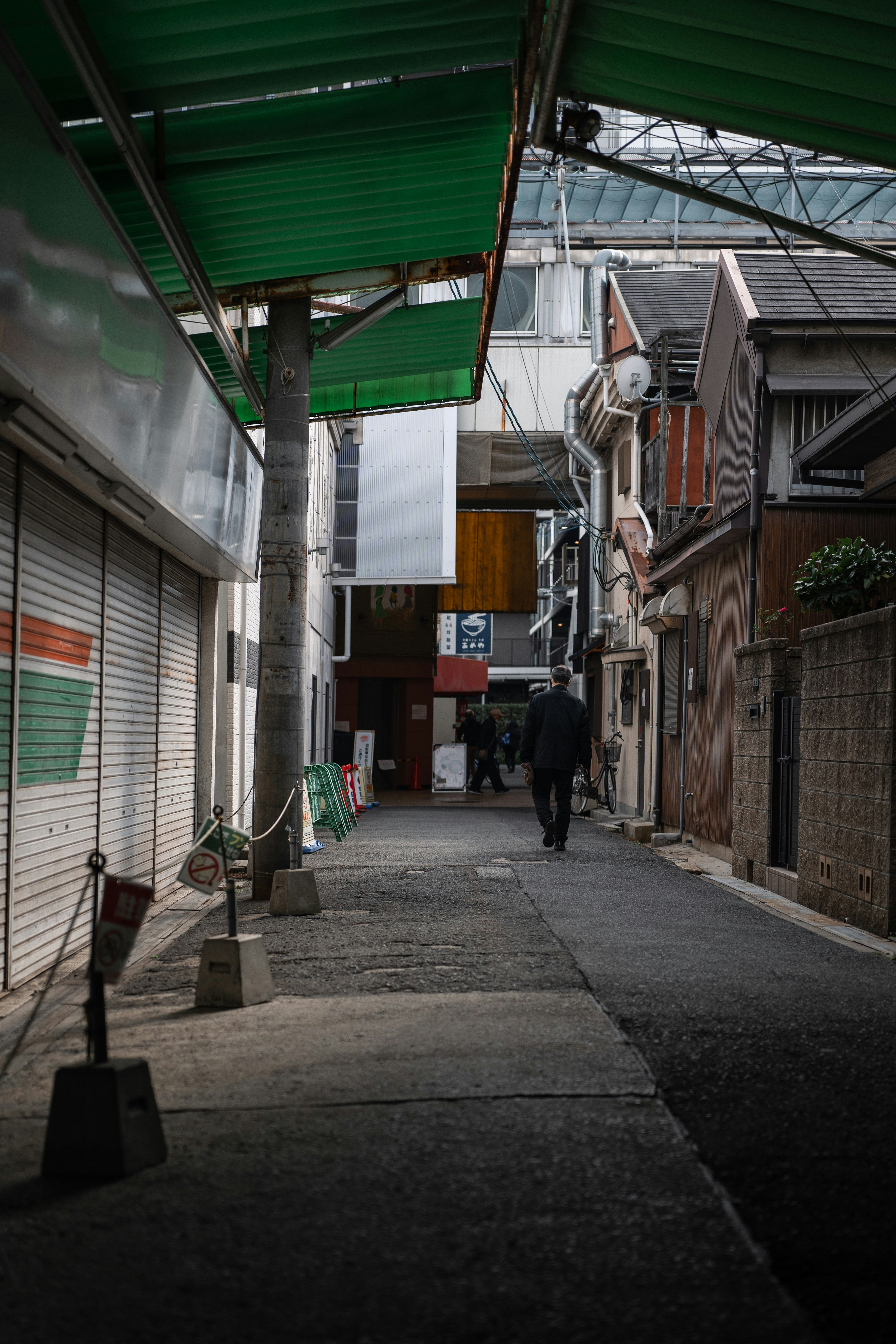 狭い路地に人が歩いている様子 緑の屋根と閉まったシャッターの店舗が見える