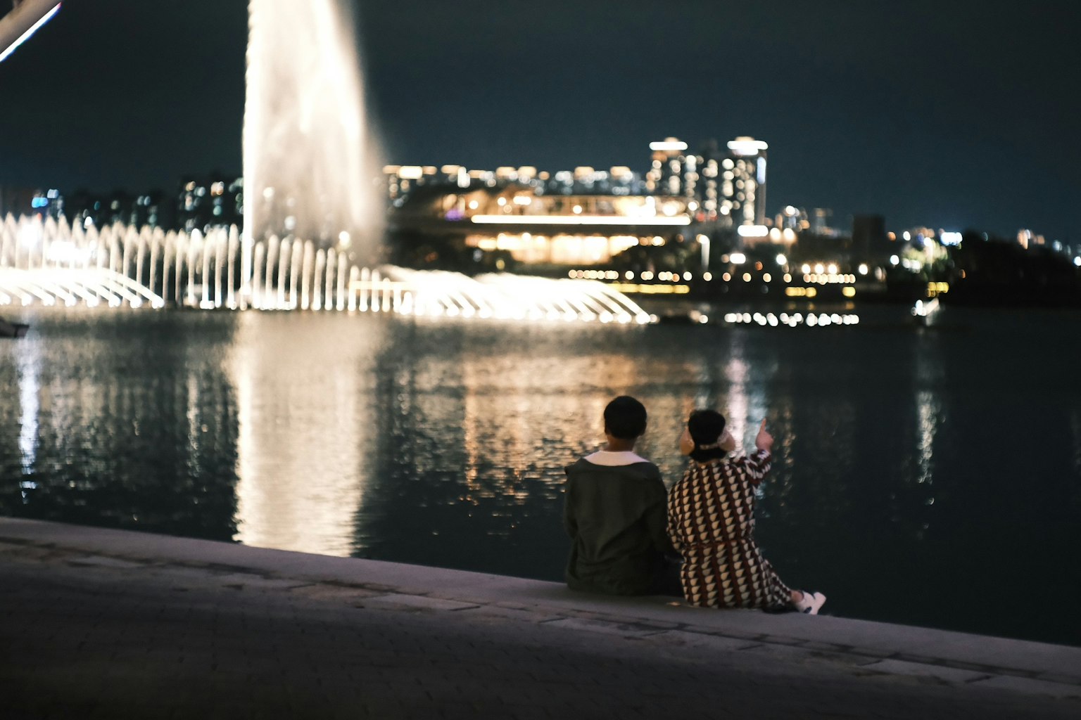 夜の水辺で座る二人の人物と噴水の光景