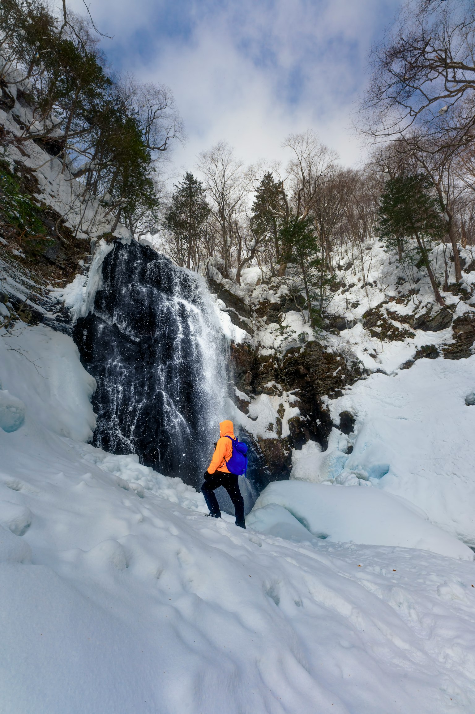Hiker in orange jacket standing near a frozen waterfall in snowy landscape