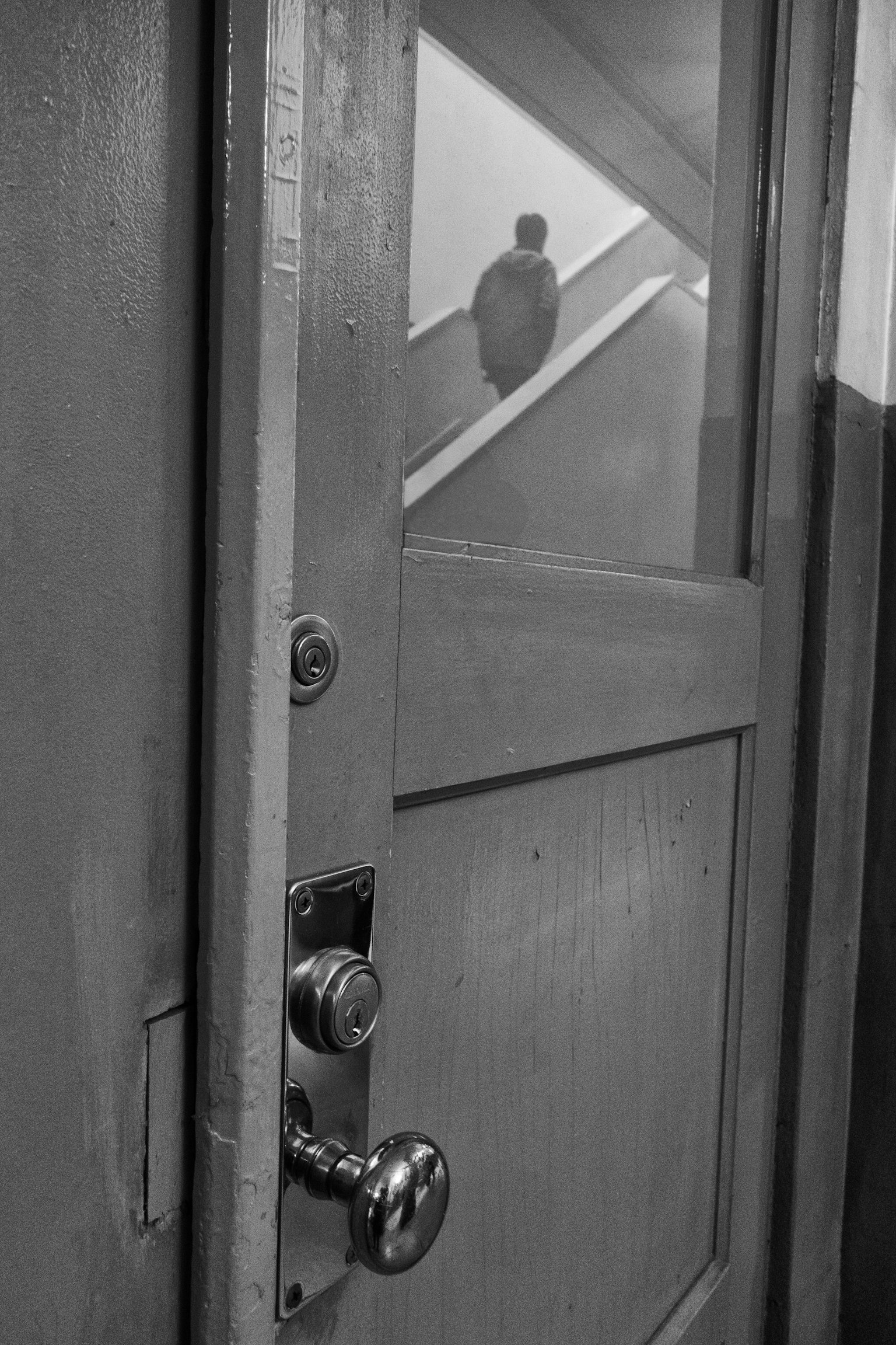 Pintu kayu dengan pegangan dan lubang kunci yang terlihat Latar belakang menampilkan tangga dan sosok bayangan