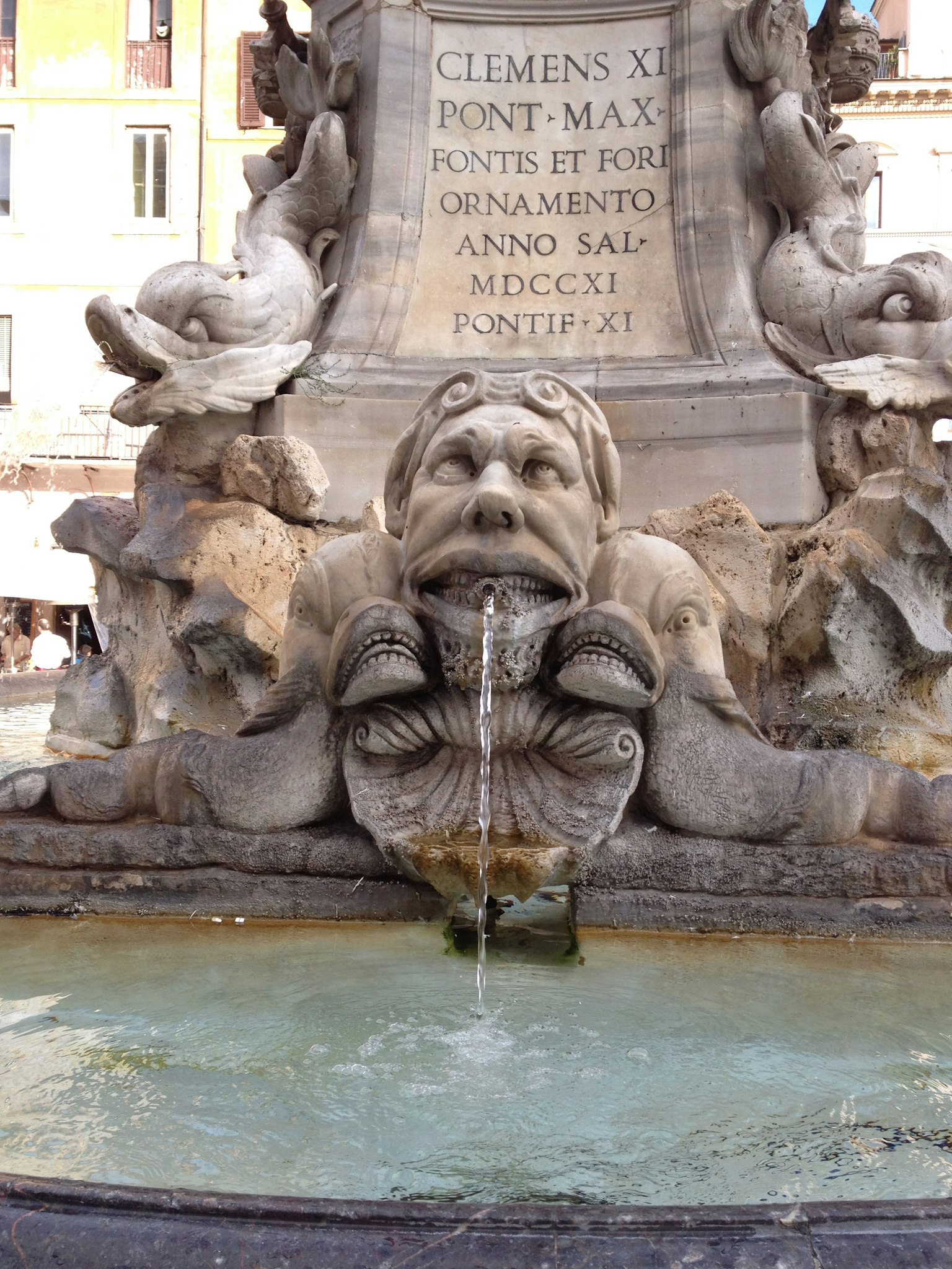A sculpted face spouting water as part of a fountain surrounded by fish sculptures