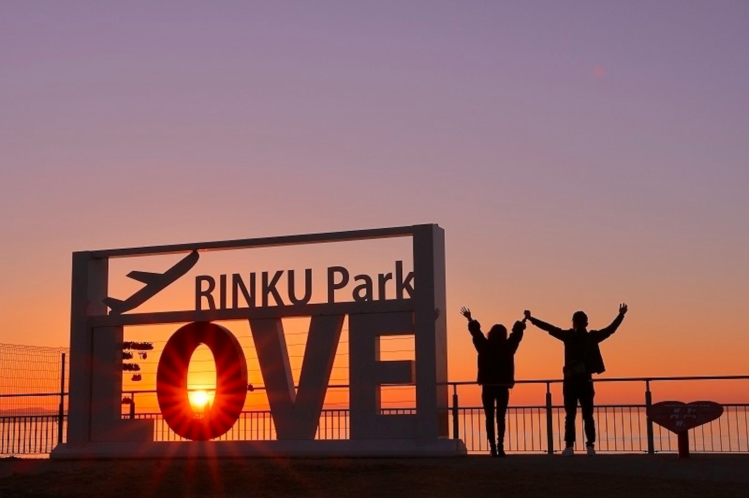 Hai người tạo dáng trước biển hiệu LOVE tại công viên RINKU với phông nền là hoàng hôn