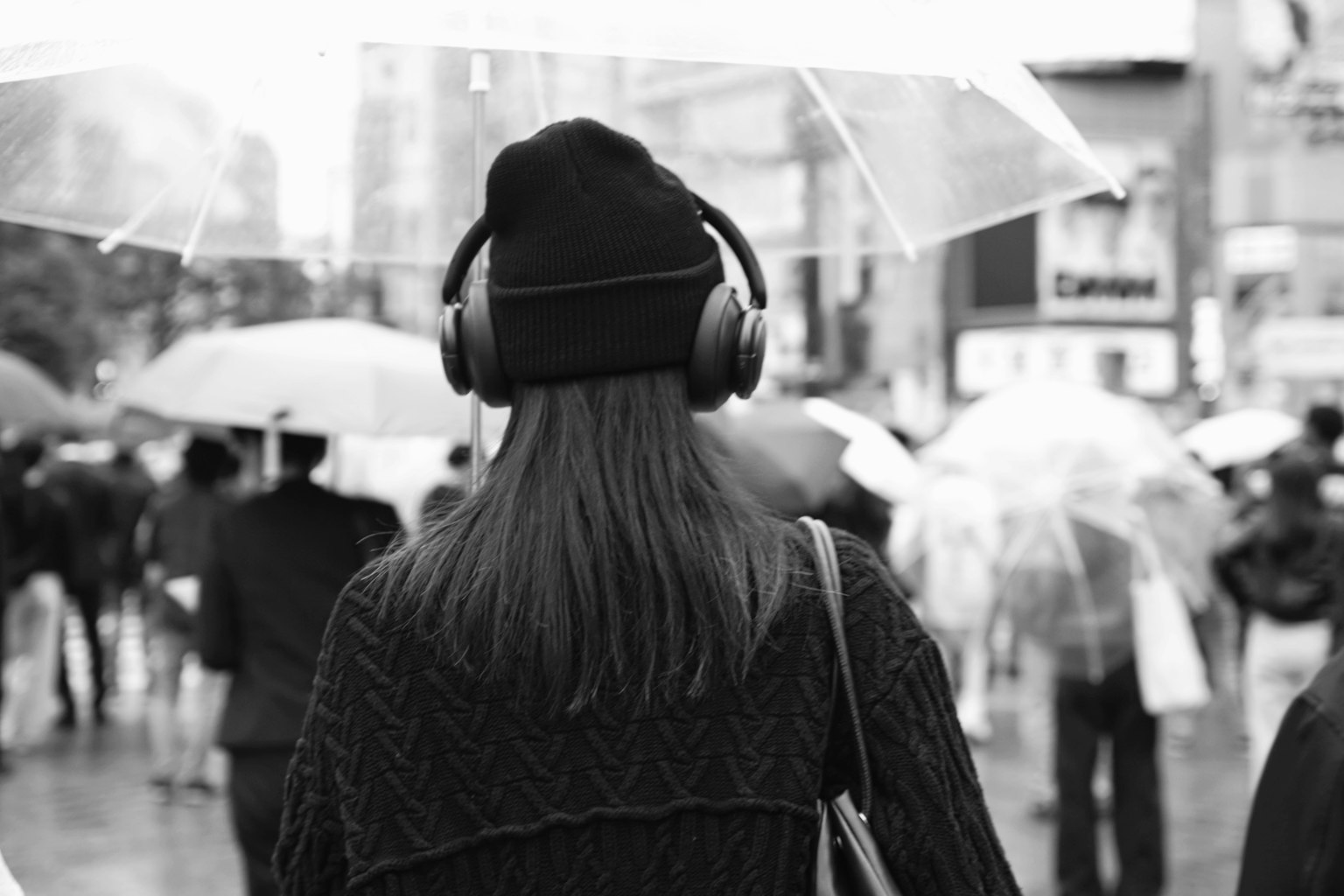 Seorang wanita mengenakan headphone berdiri membelakangi kamera dalam pemandangan hitam putih yang hujan