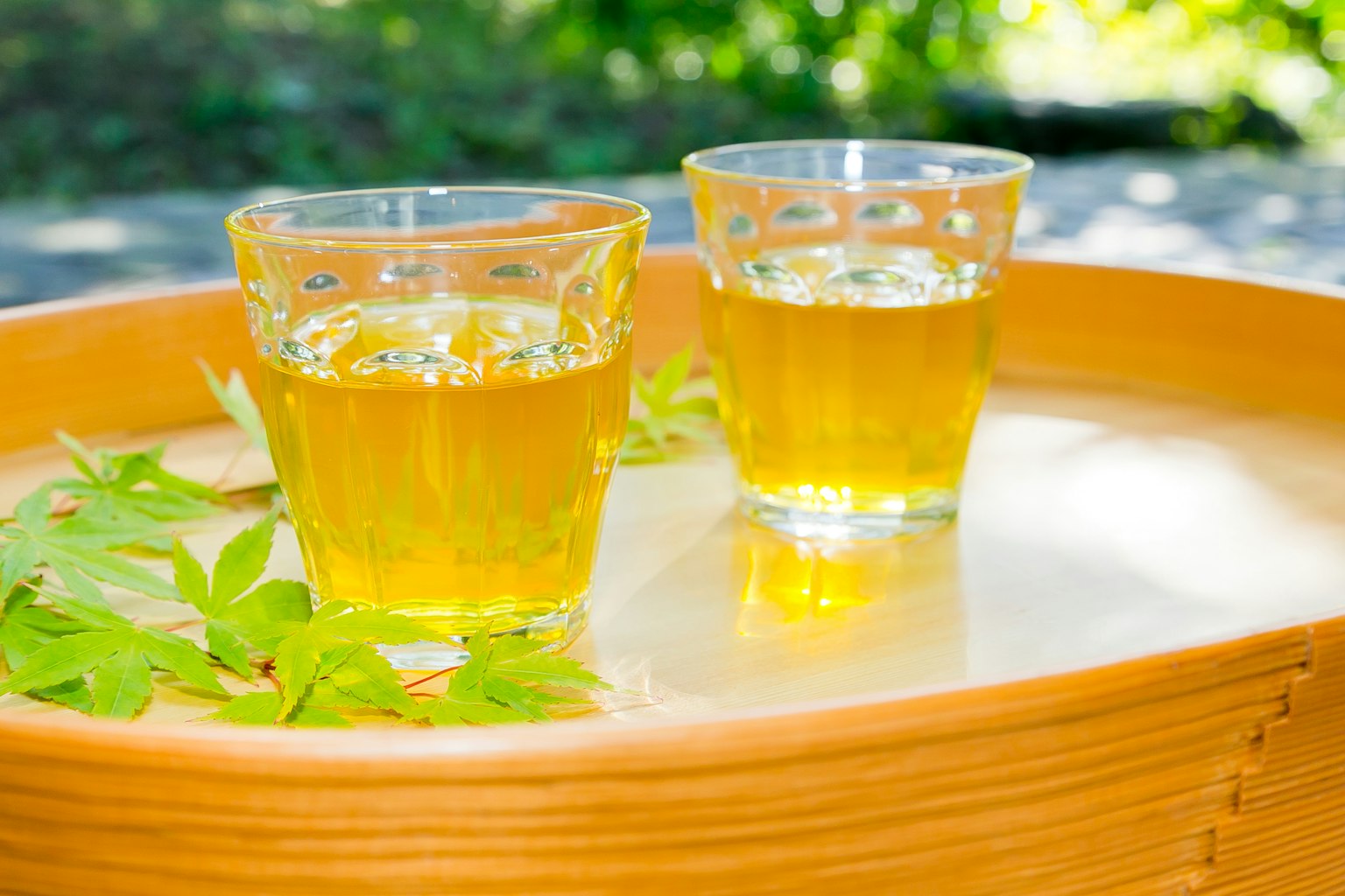 Zwei transparente Gläser mit gelbem Getränk auf einem Holztablett umgeben von grünen Blättern