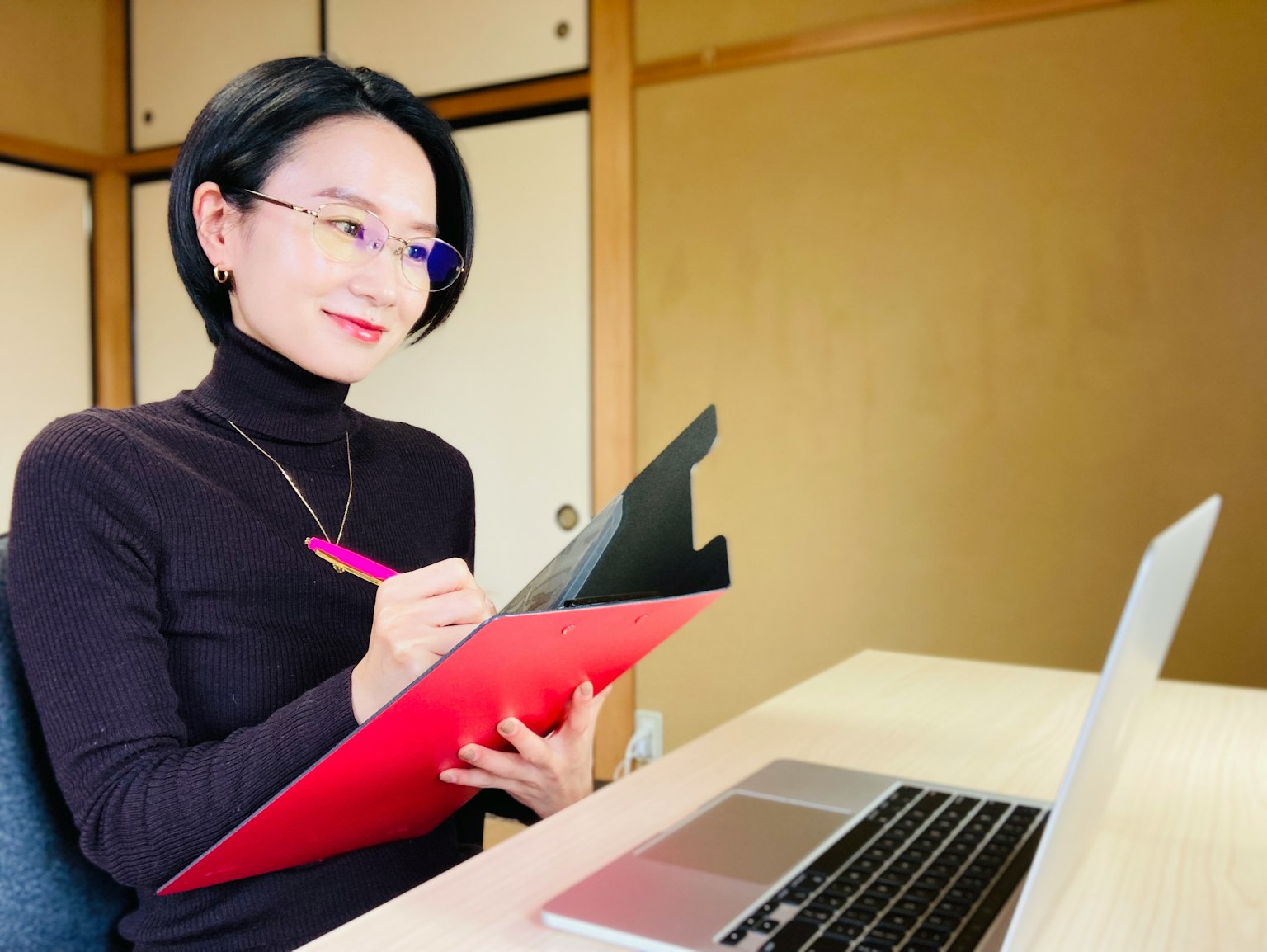 Femme assise devant un ordinateur portable dans un bureau moderne prenant des notes dans un dossier rouge