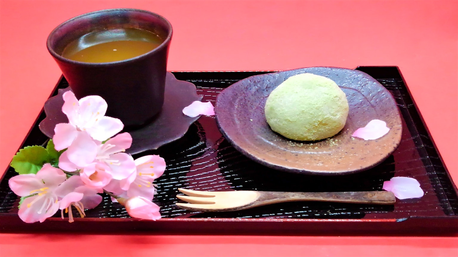 A green Japanese sweet and tea served on a black plate and tray with cherry blossom petals