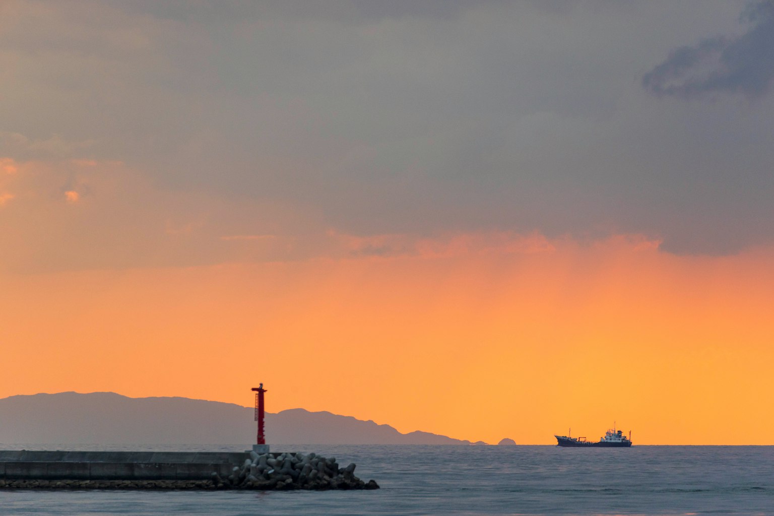 Lighthouse standing on the sea at sunset with a distant ship