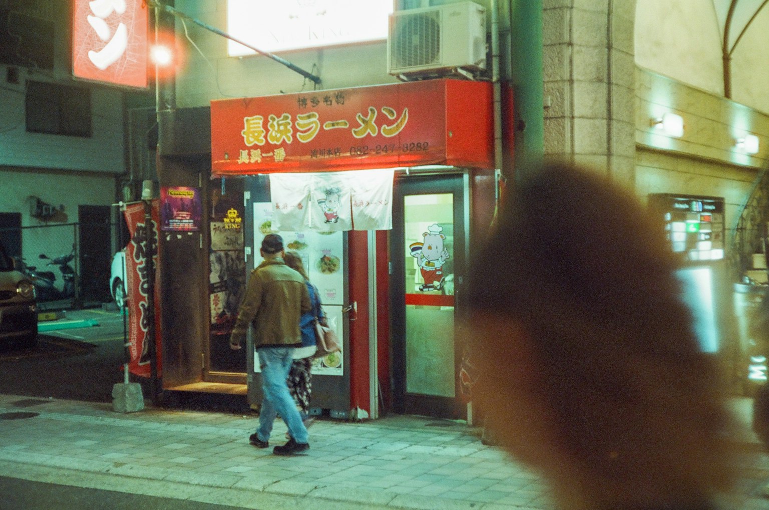 Cảnh đêm của một quán ramen với biển hiệu màu đỏ và người đi qua