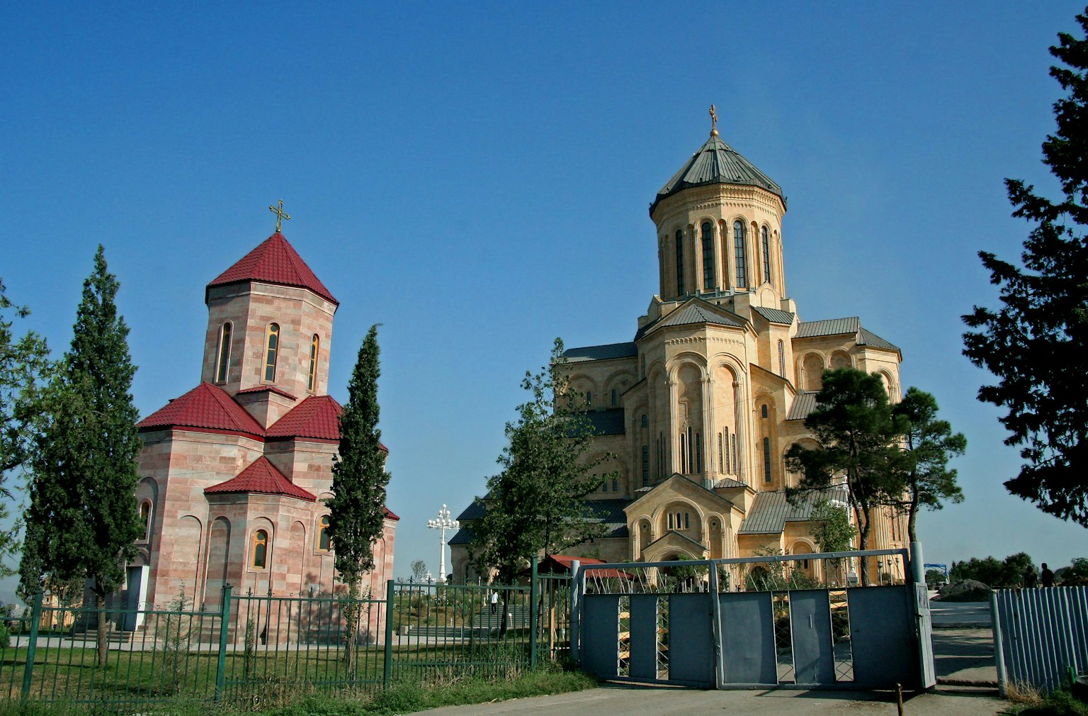 Dos edificios de iglesia bajo un cielo azul rodeados de árboles verdes con una pequeña iglesia de techo rojo y una gran iglesia dorada