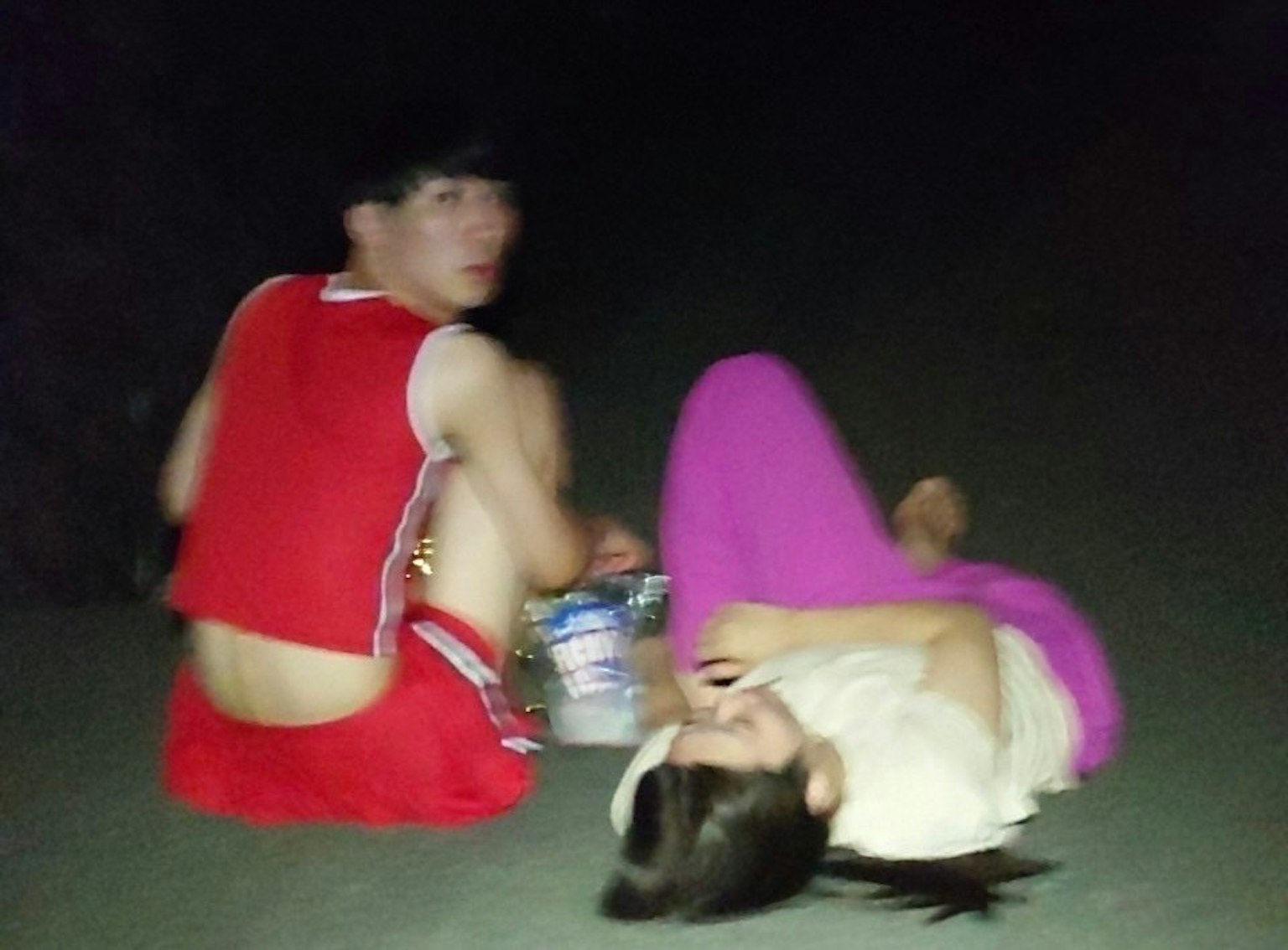 Dua pemuda duduk di malam hari satu mengenakan kaos merah memegang camilan