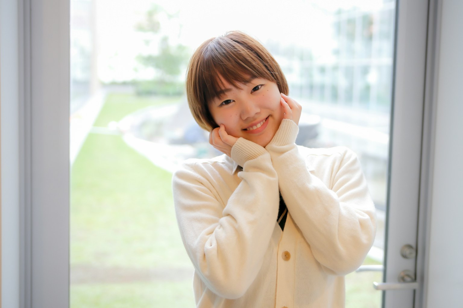 Eine lächelnde Frau in einem weißen Pullover steht am Fenster