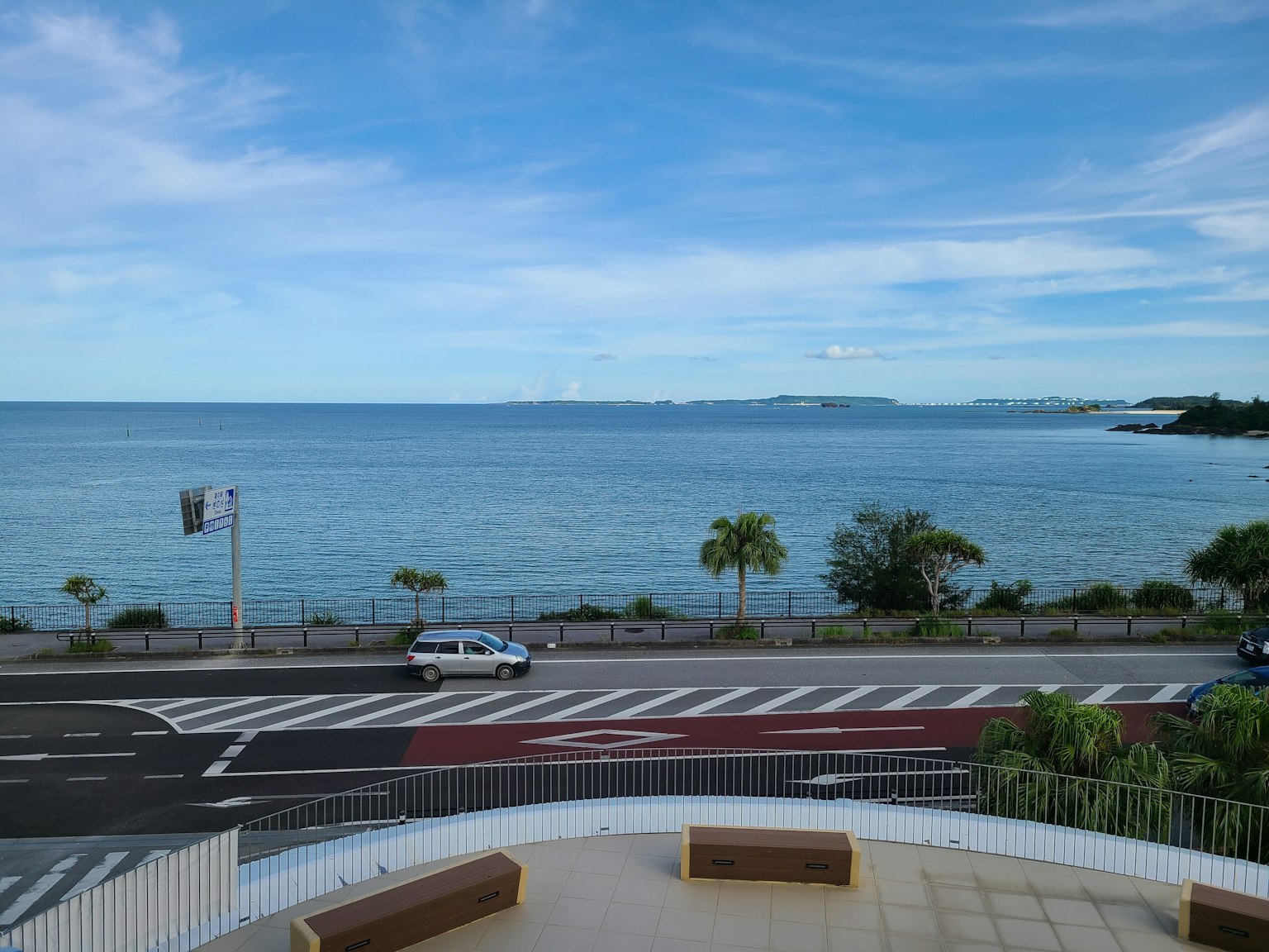 Scenic view of blue ocean and sky with a road featuring a car and palm trees