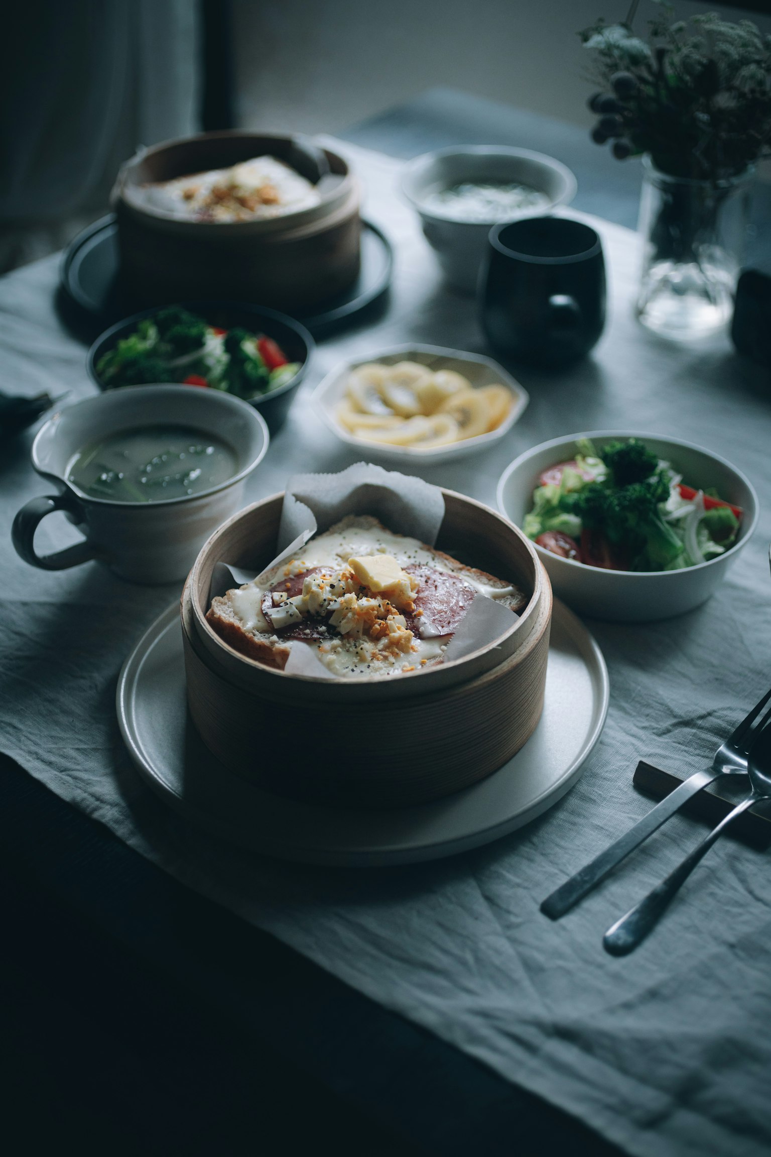 Ein Esstisch mit Gerichten einschließlich eines Dampfgarers voller Speisen und Salate neben Tassen und Besteck