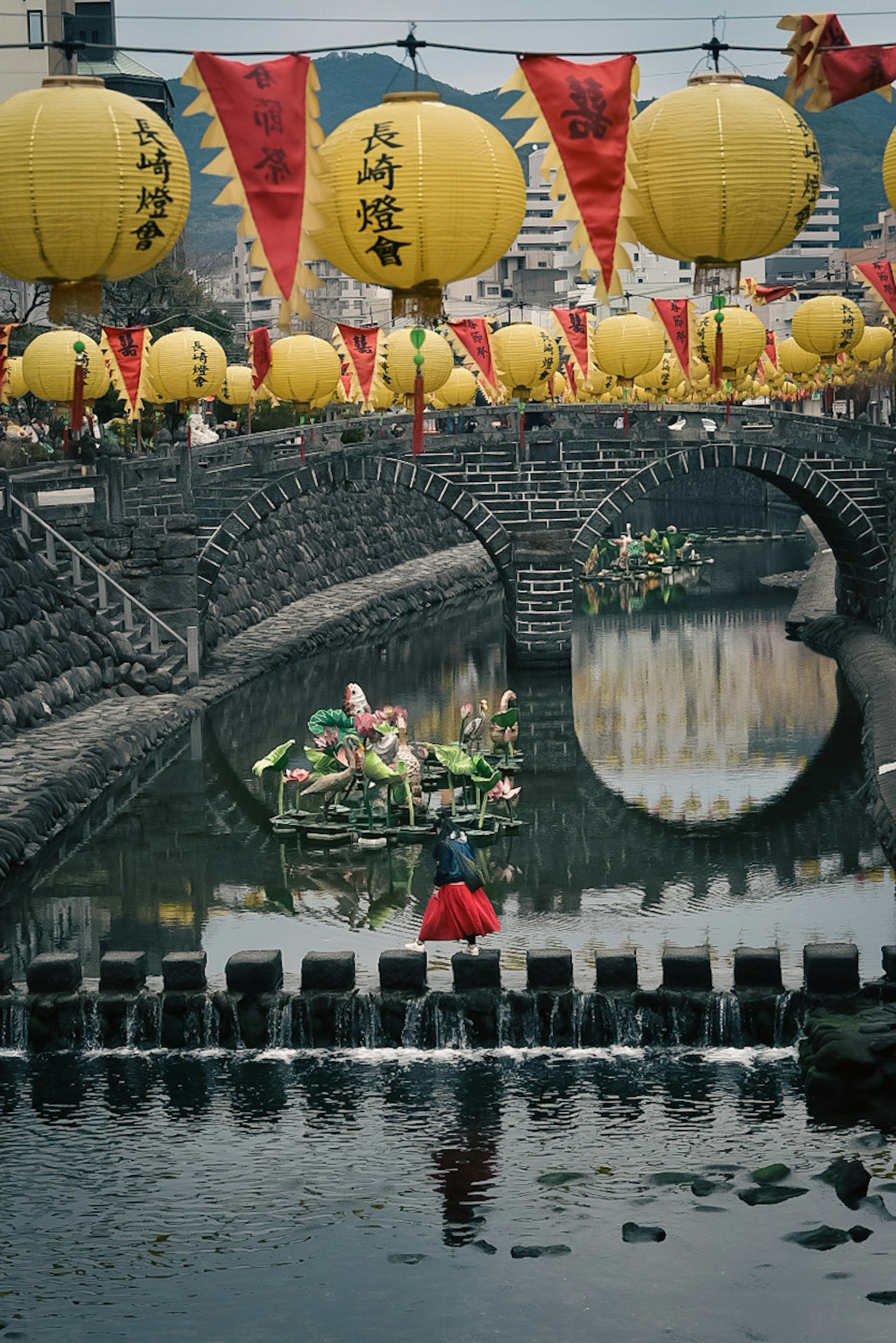 A scenic view of colorful lanterns and flags above a stone bridge with people in boats on a calm river