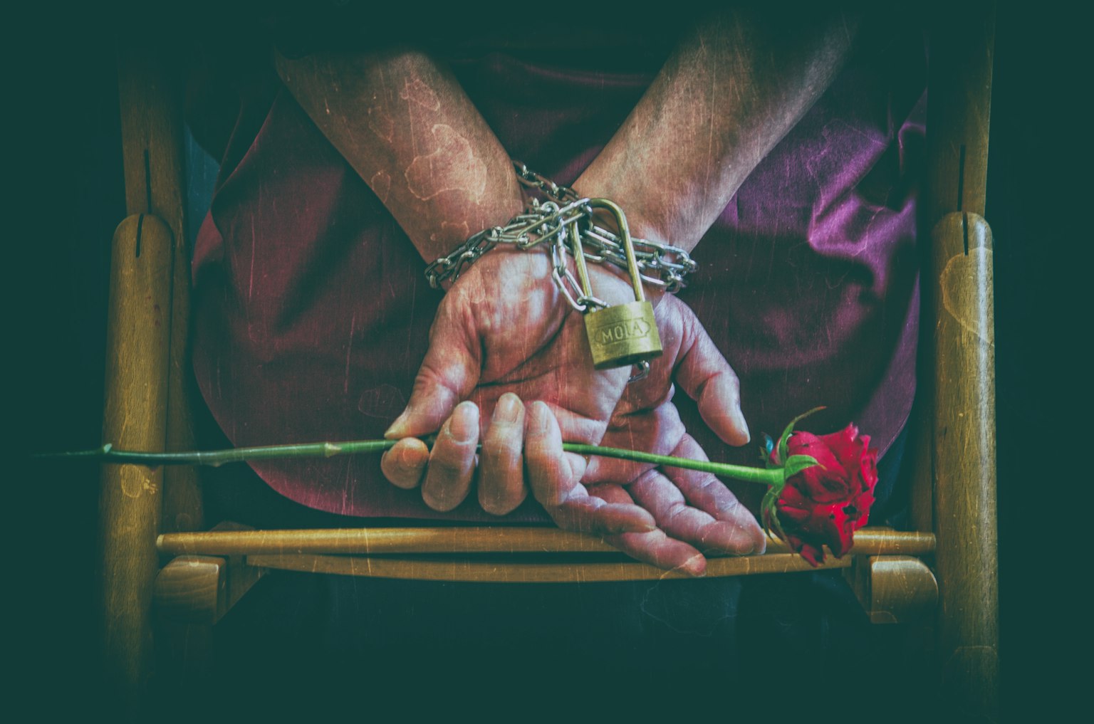 Mani di una persona ammanettate con un lucchetto che tengono una rosa rossa dietro la schiena
