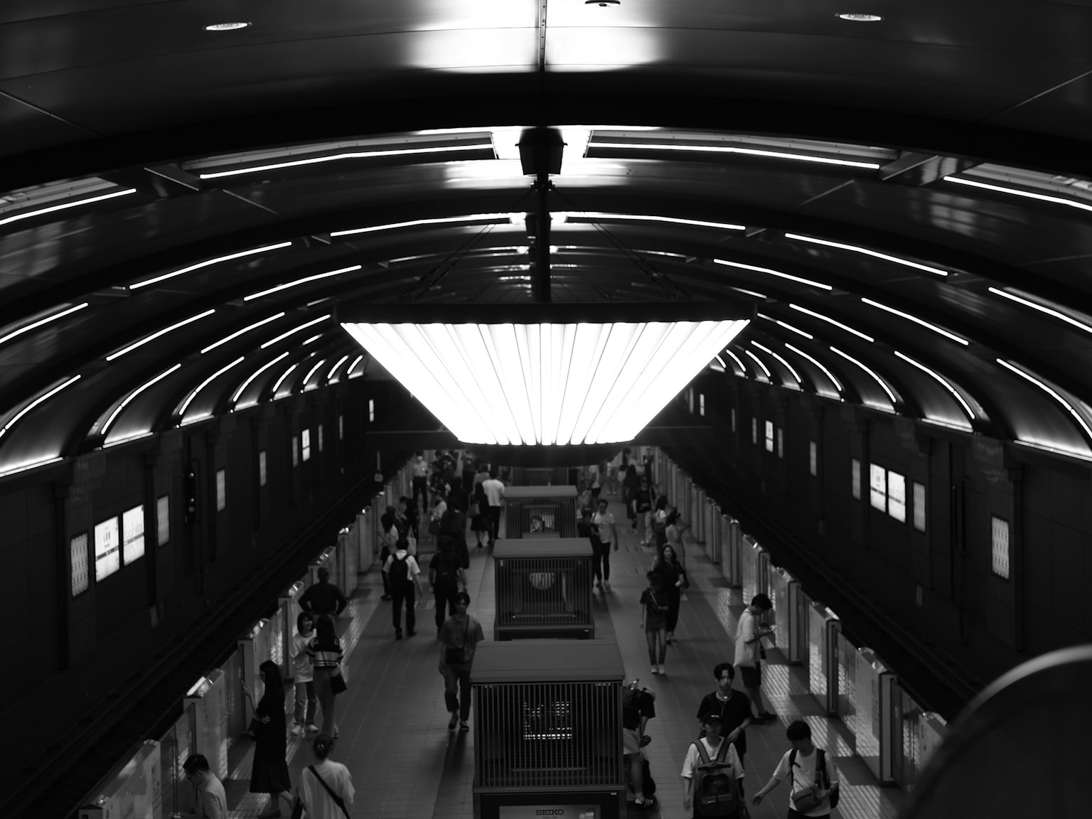 Nội thất đen trắng của một ga tàu điện ngầm với mọi người đi lại dưới ánh đèn trần hình vòm đặc trưng