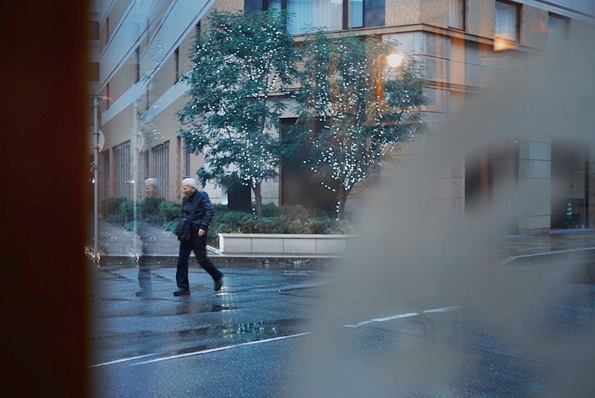 雨上がりの街角で歩く人とガラス越しの木々「画像」