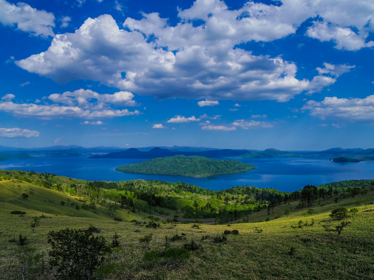 Malersiche Aussicht auf einen See umgeben von grünen Hügeln und Inseln unter einem blauen Himmel mit flauschigen Wolken