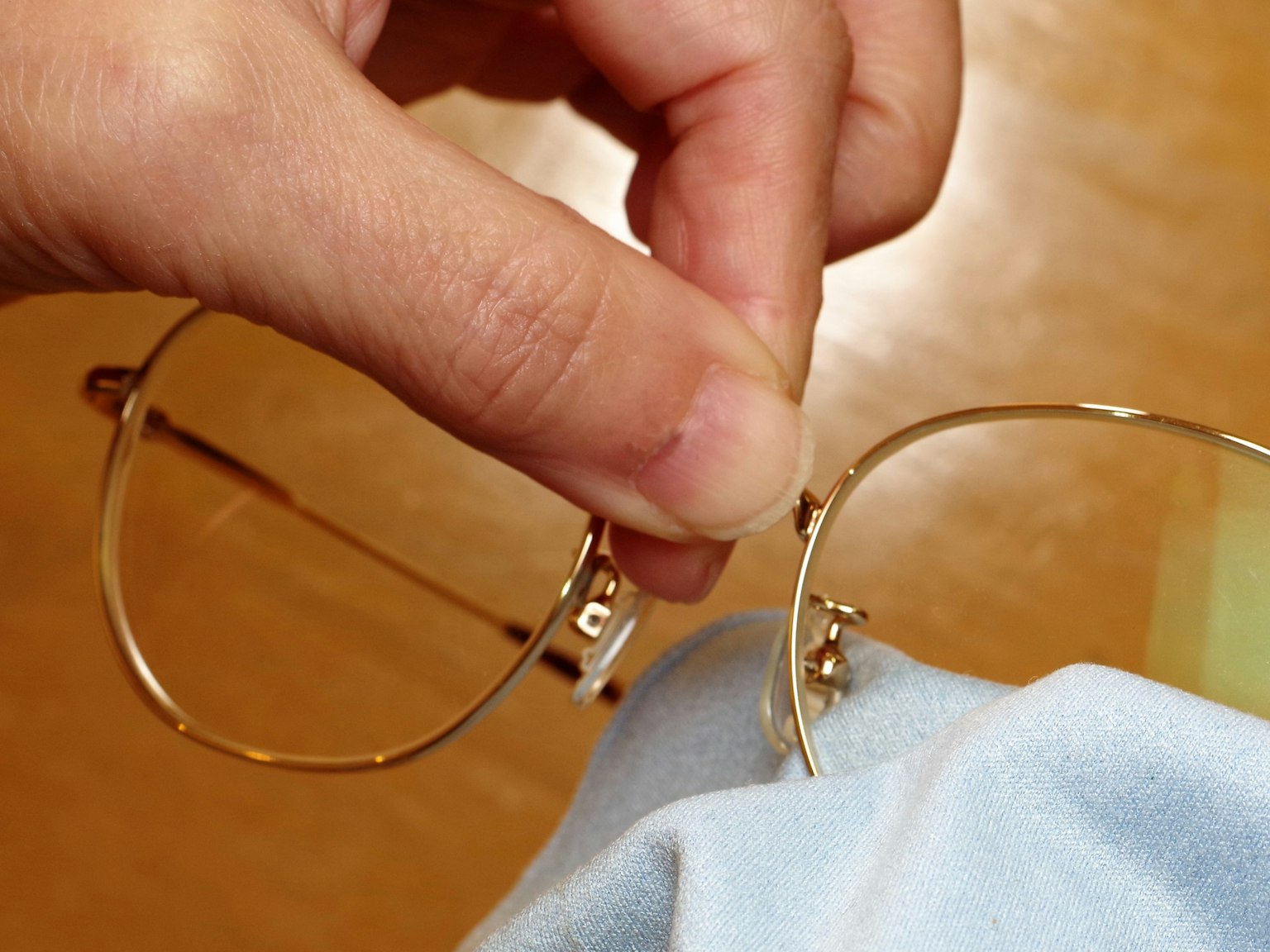 Eine Hand hält eine Brille mit einem goldenen Rahmen, die mit einem blauen Tuch gereinigt wird