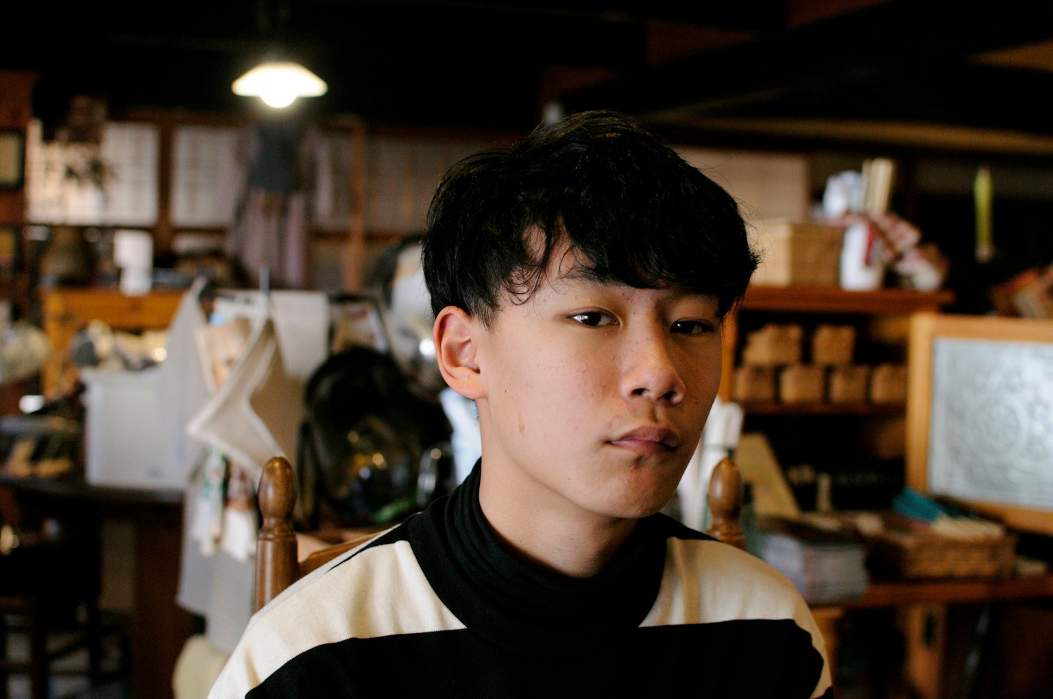 若い男性がカジュアルな服装で座っている背景には木製の家具と雑多なアイテムが見える