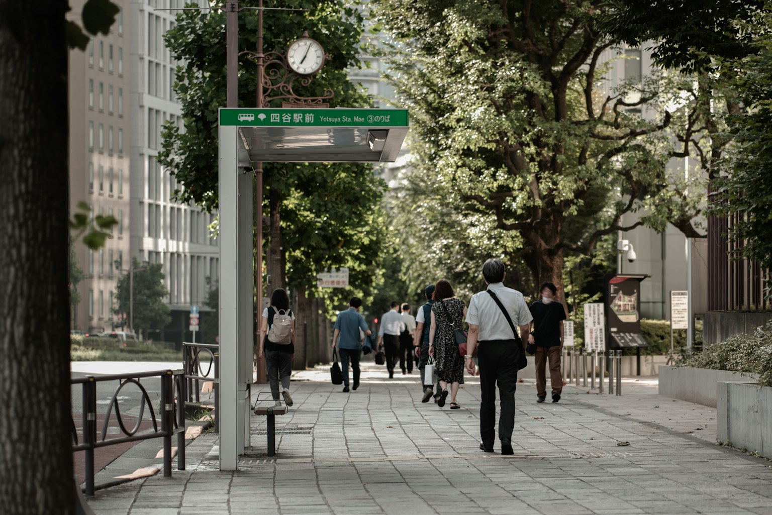 Personas caminando por una calle arbolada con una parada de autobús y un reloj