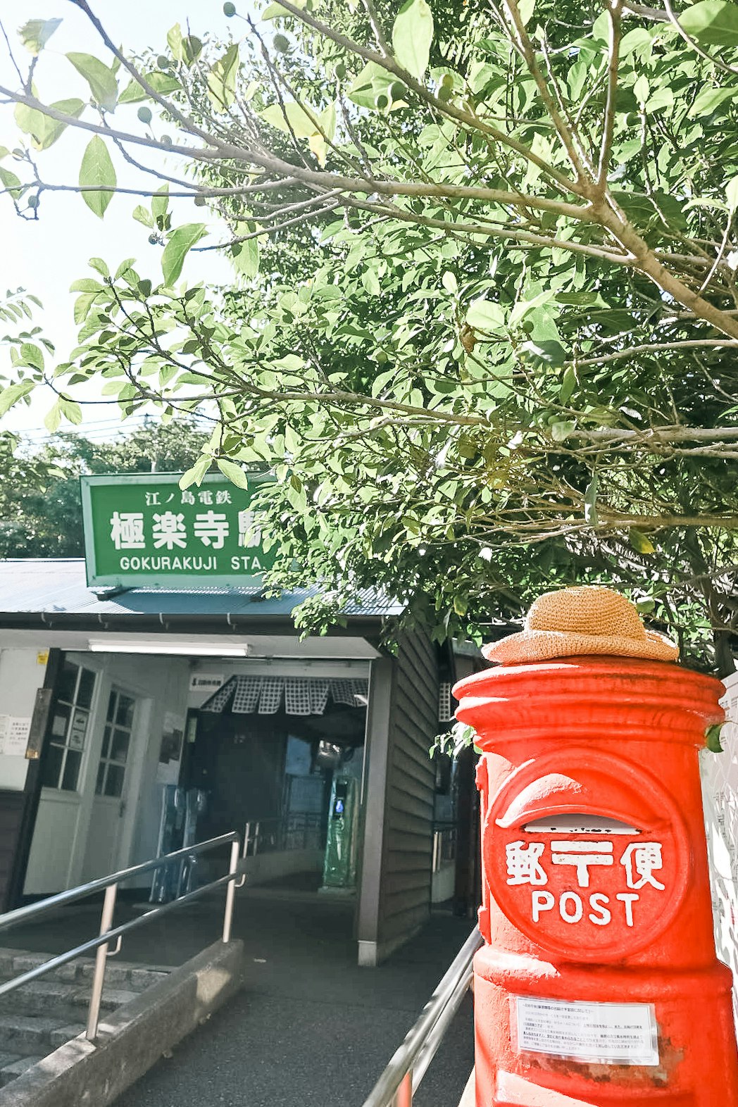 緑の木々の下にある駅入口と赤い郵便ポストのある風景