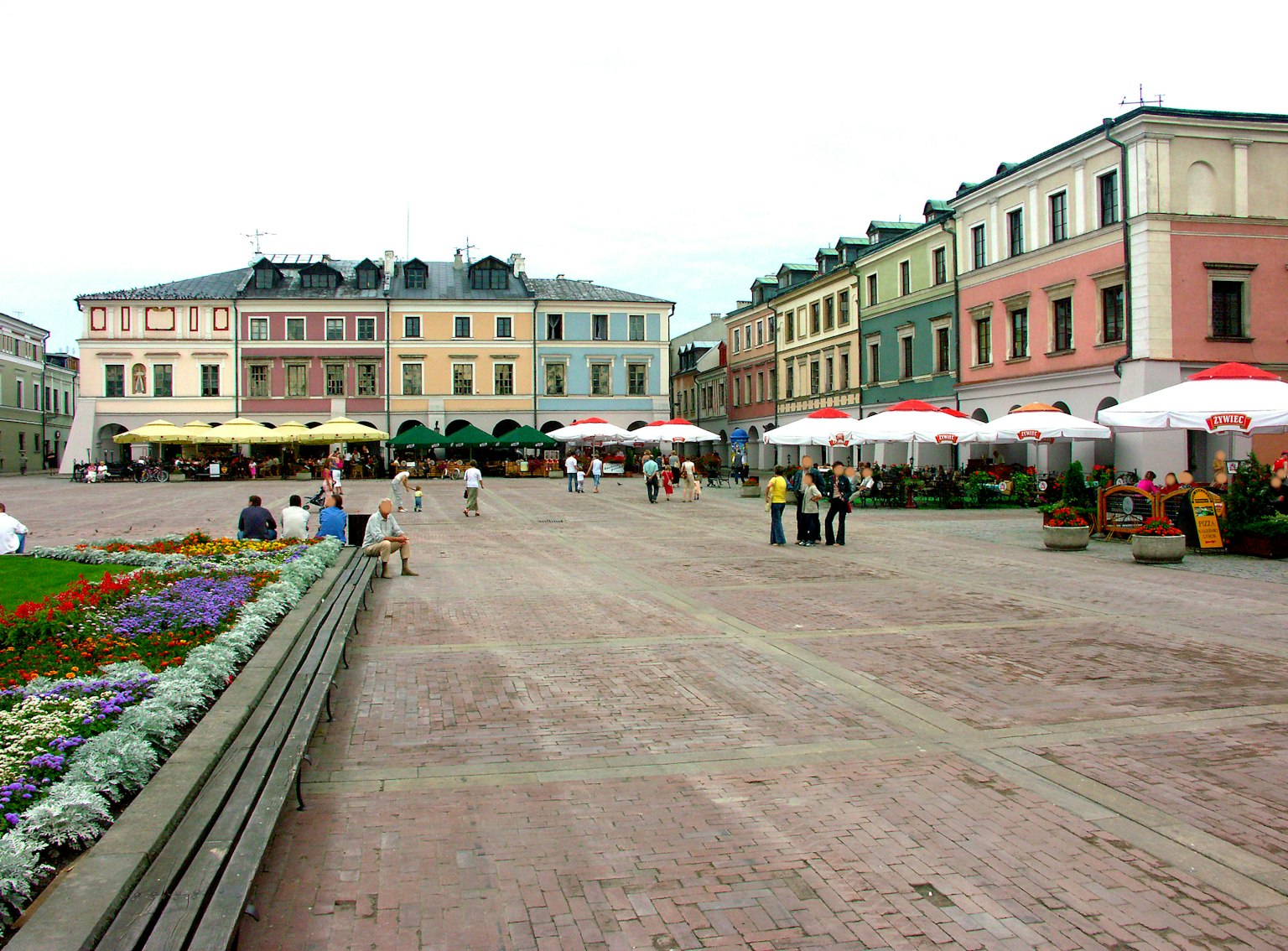 Vista de una plaza colorida con edificios y cafeterías al aire libre con sombrillas y parterres de flores