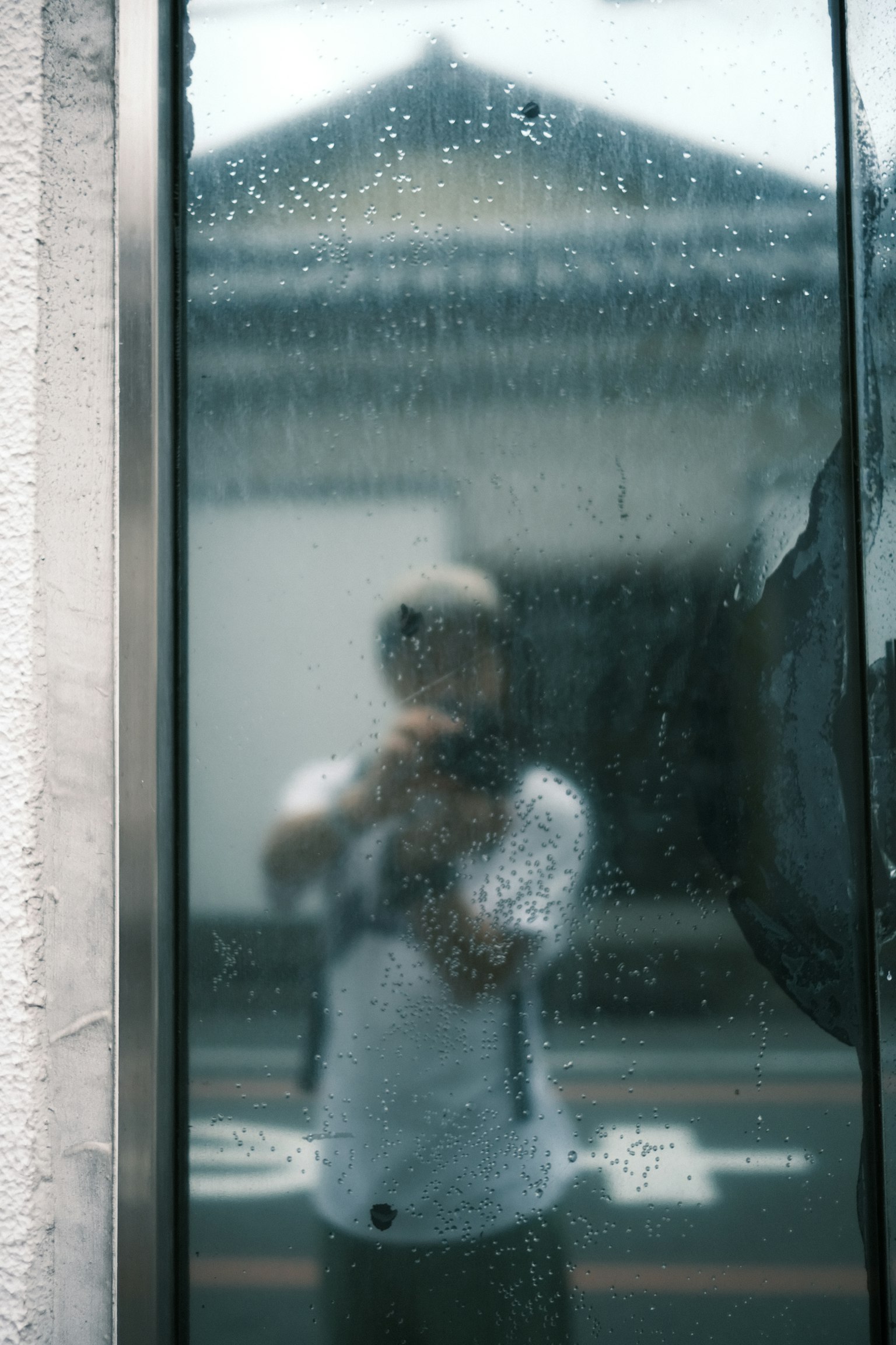 Persona tomando un selfie reflejada en un vidrio cubierto de lluvia con un edificio tradicional al fondo