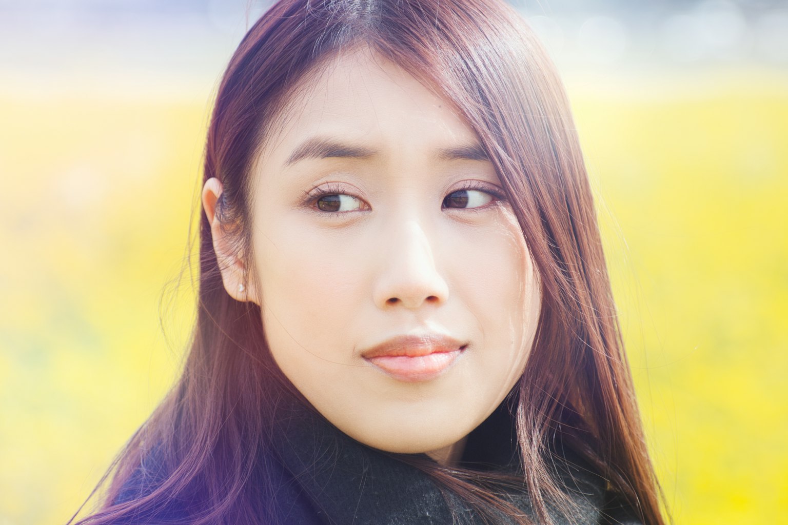 A young woman gazes thoughtfully with a blurred yellow flower field in the background
