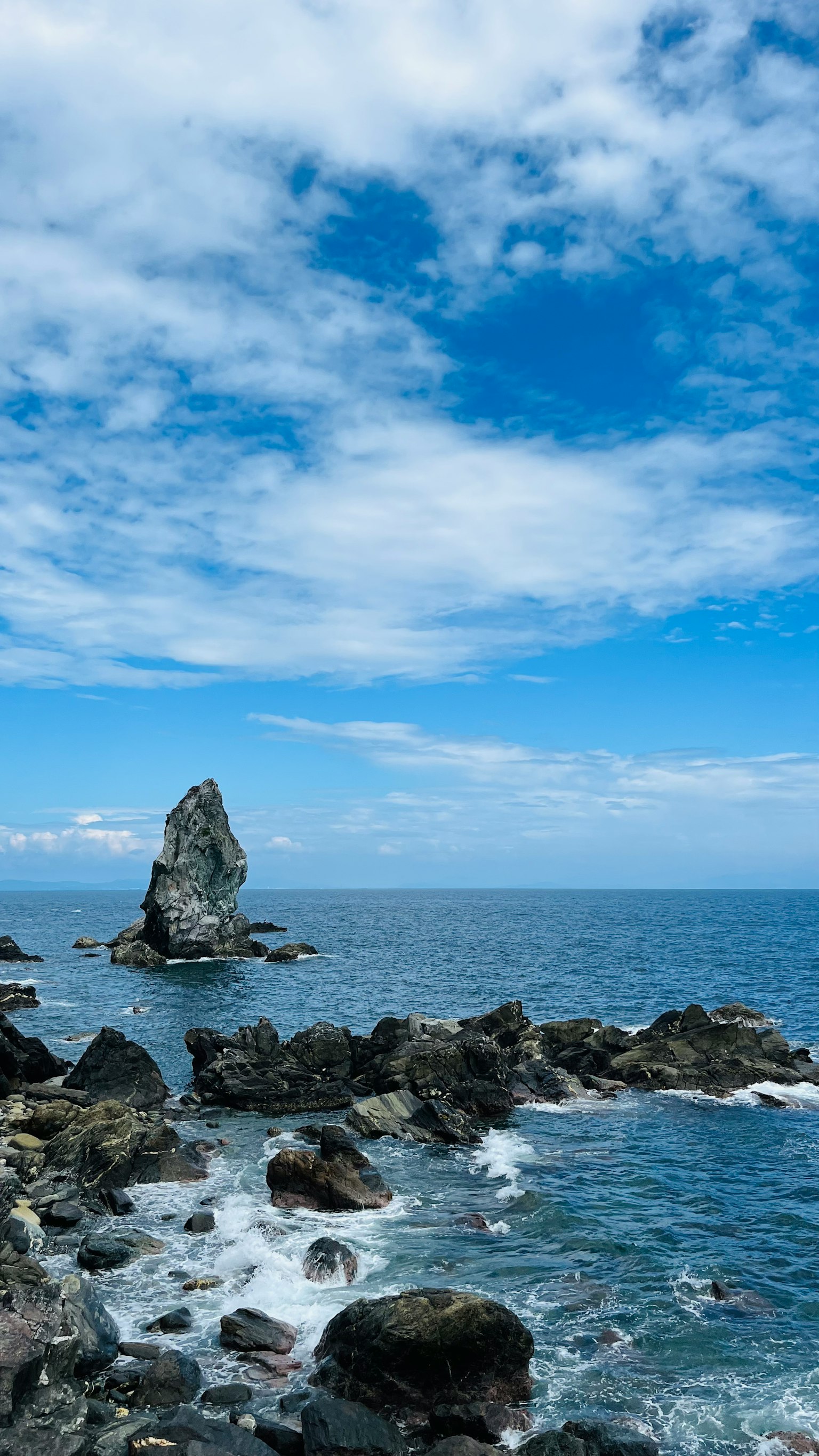 A tall rock standing in the ocean with waves crashing against a rocky shore under a blue sky