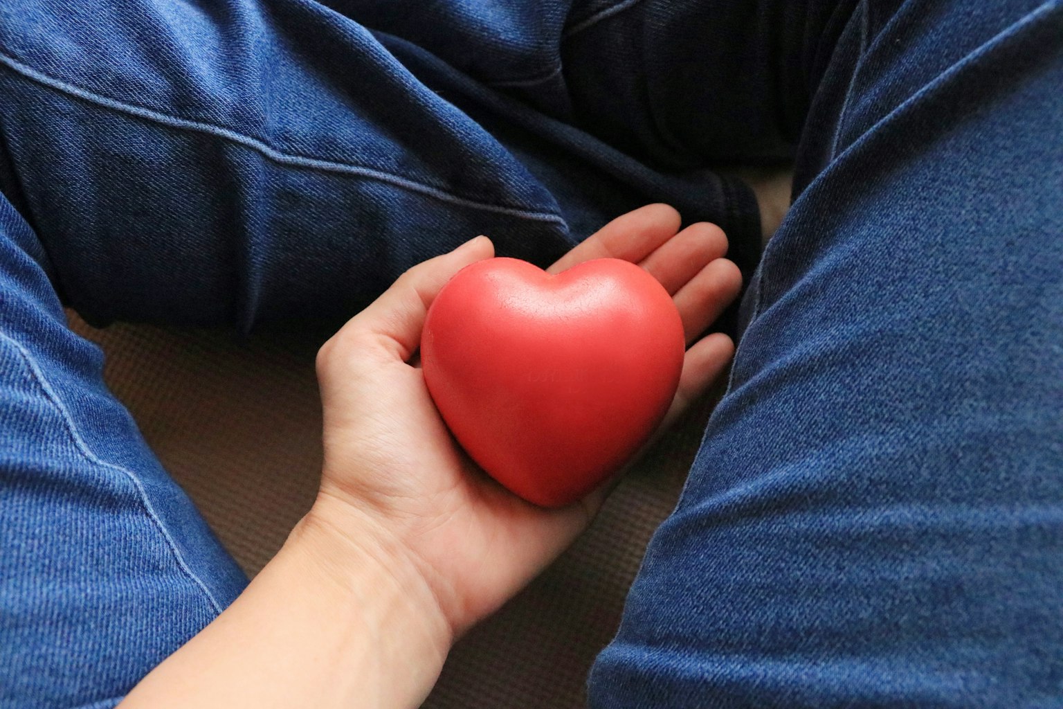 A hand holding a red heart-shaped object with denim jeans visible