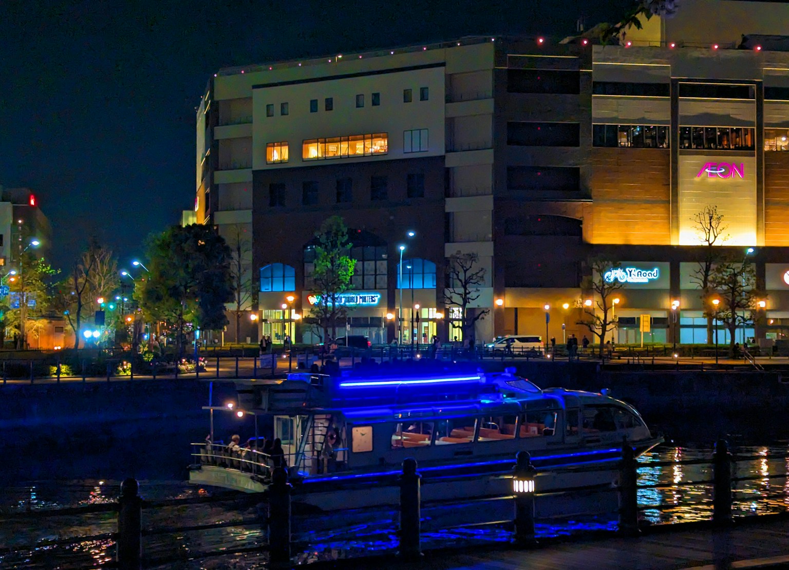 เรือที่มีแสงสีน้ำเงินลอยอยู่บนแม่น้ำในเวลากลางคืนพร้อมอาคารที่มีแสงสว่าง