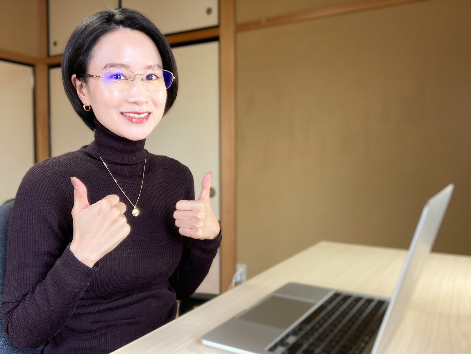 Eine Frau in einem schwarzen Rollkragenpullover zeigt Daumen hoch, während sie an einem Schreibtisch mit einem Laptop sitzt