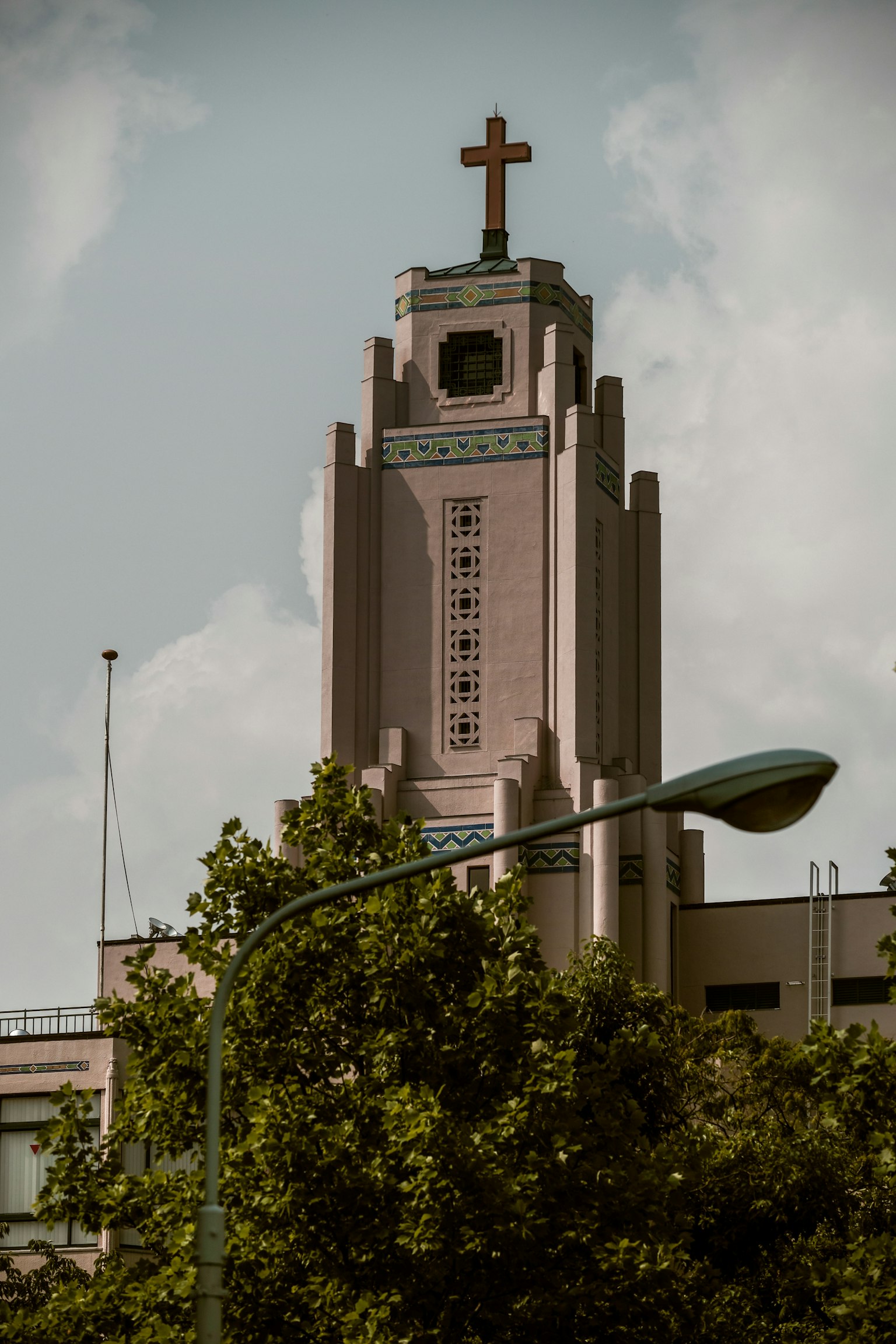 Foto eines Gebäudes mit einem hohen Turm und einem Kreuz oben Grüne Bäume und eine Straßenlaterne im Vordergrund