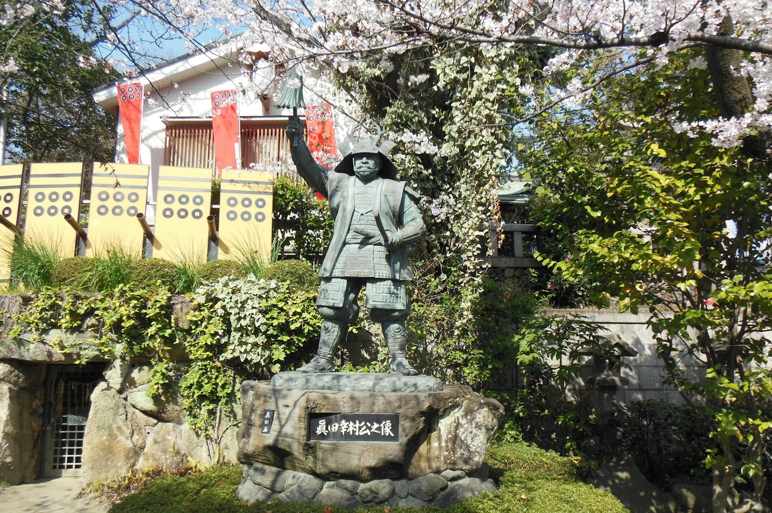 Estatua de un samurái bajo un árbol de cerezo con un edificio tradicional al fondo