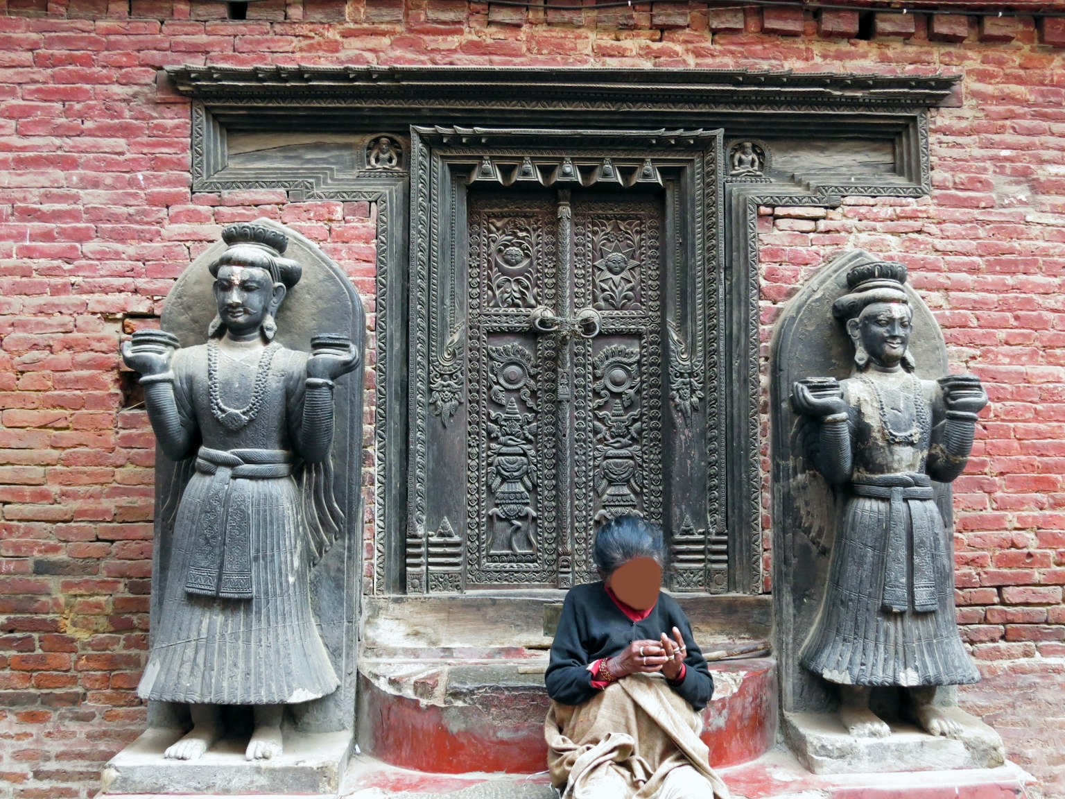 Dos estatuas de piedra con tallas intrincadas al lado de una puerta antigua con diseños detallados y una persona sentada frente a ella